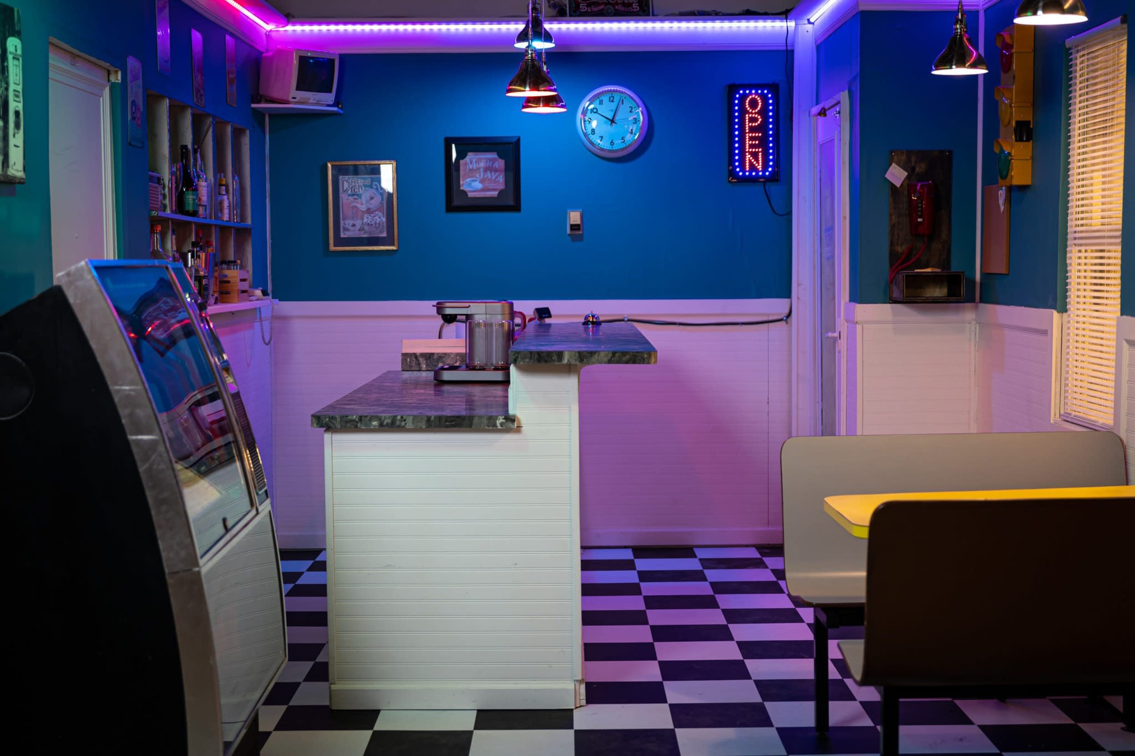 A retro diner interior with a checkered floor, a countertop, a vintage jukebox, and neon lighting.