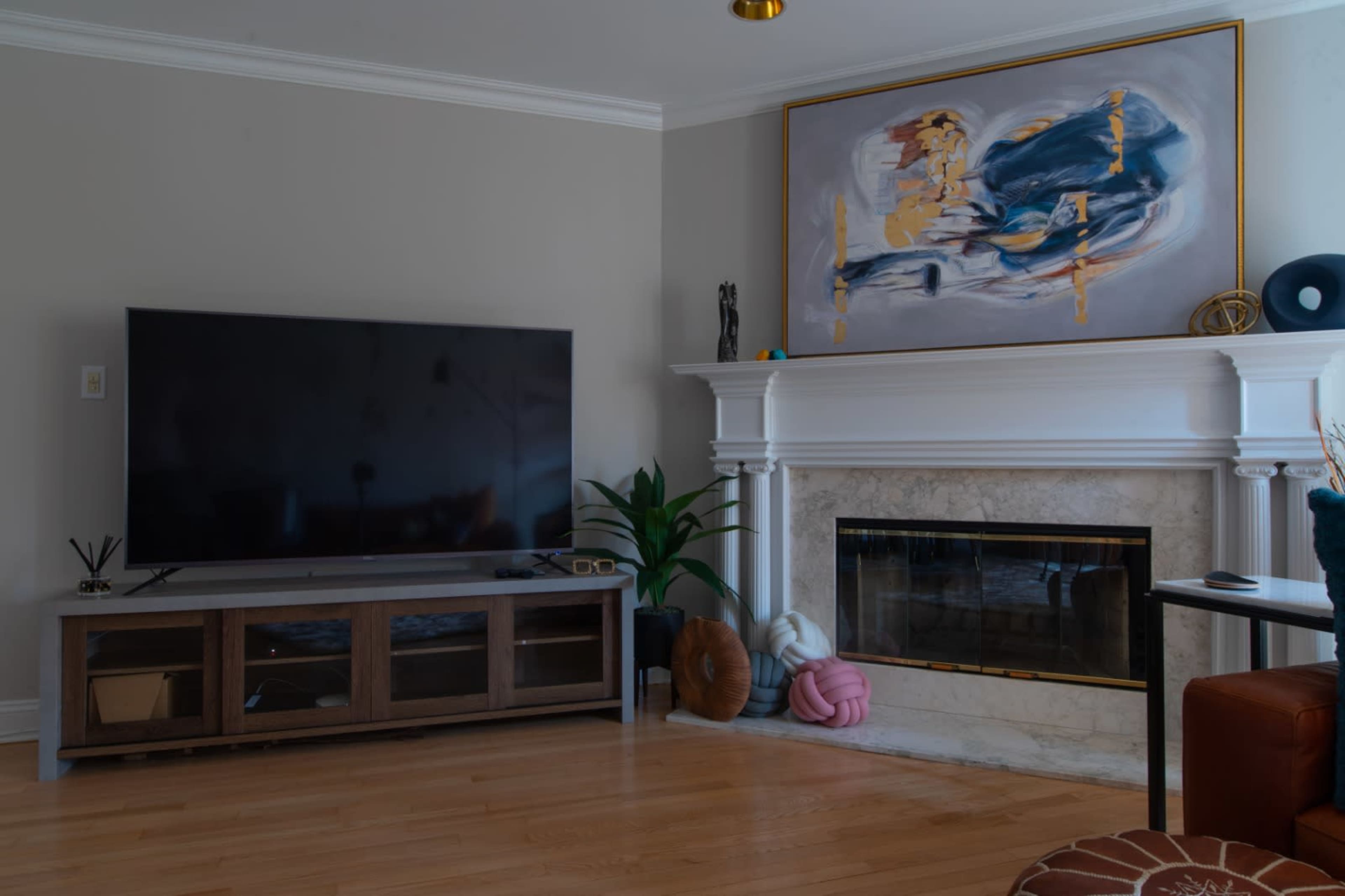 The scene features a living room with a large flat-screen TV on a wooden stand, a marble fireplace framed by a piece of abstract artwork, and decorative elements like a plant and patterned cushions.