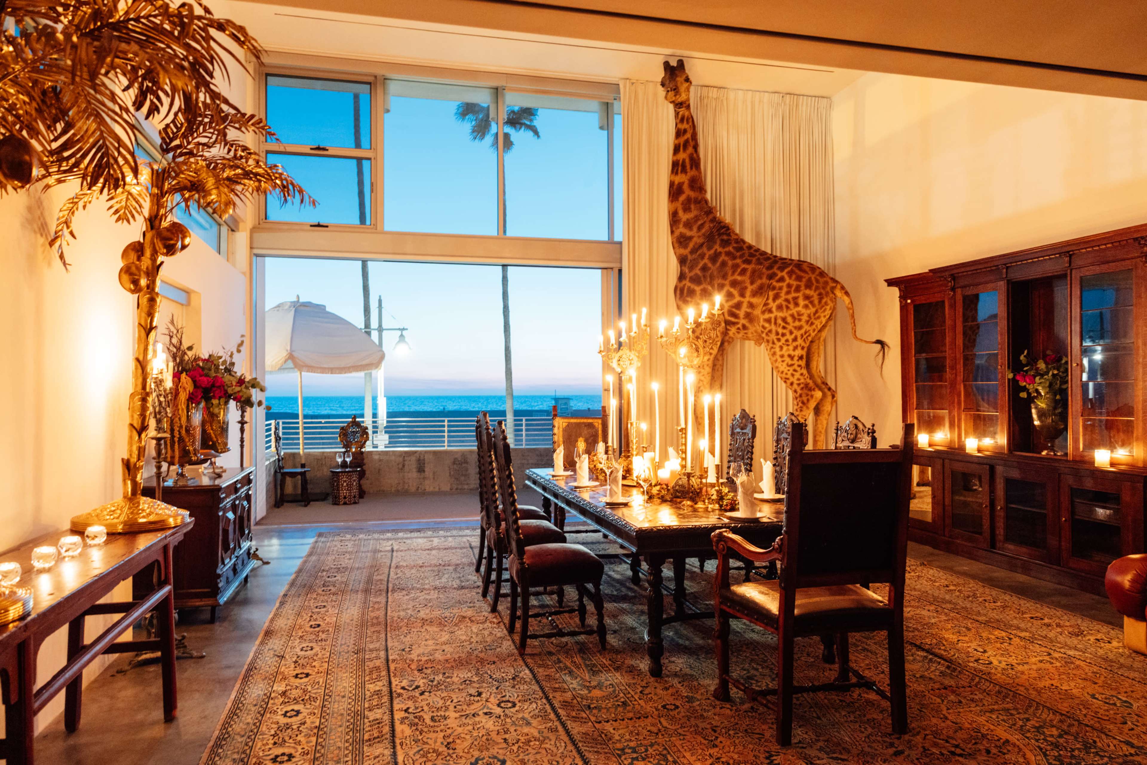 The room features a dining table set with candles, an ornate rug, and a giraffe standing by a large window overlooking the ocean at sunset.