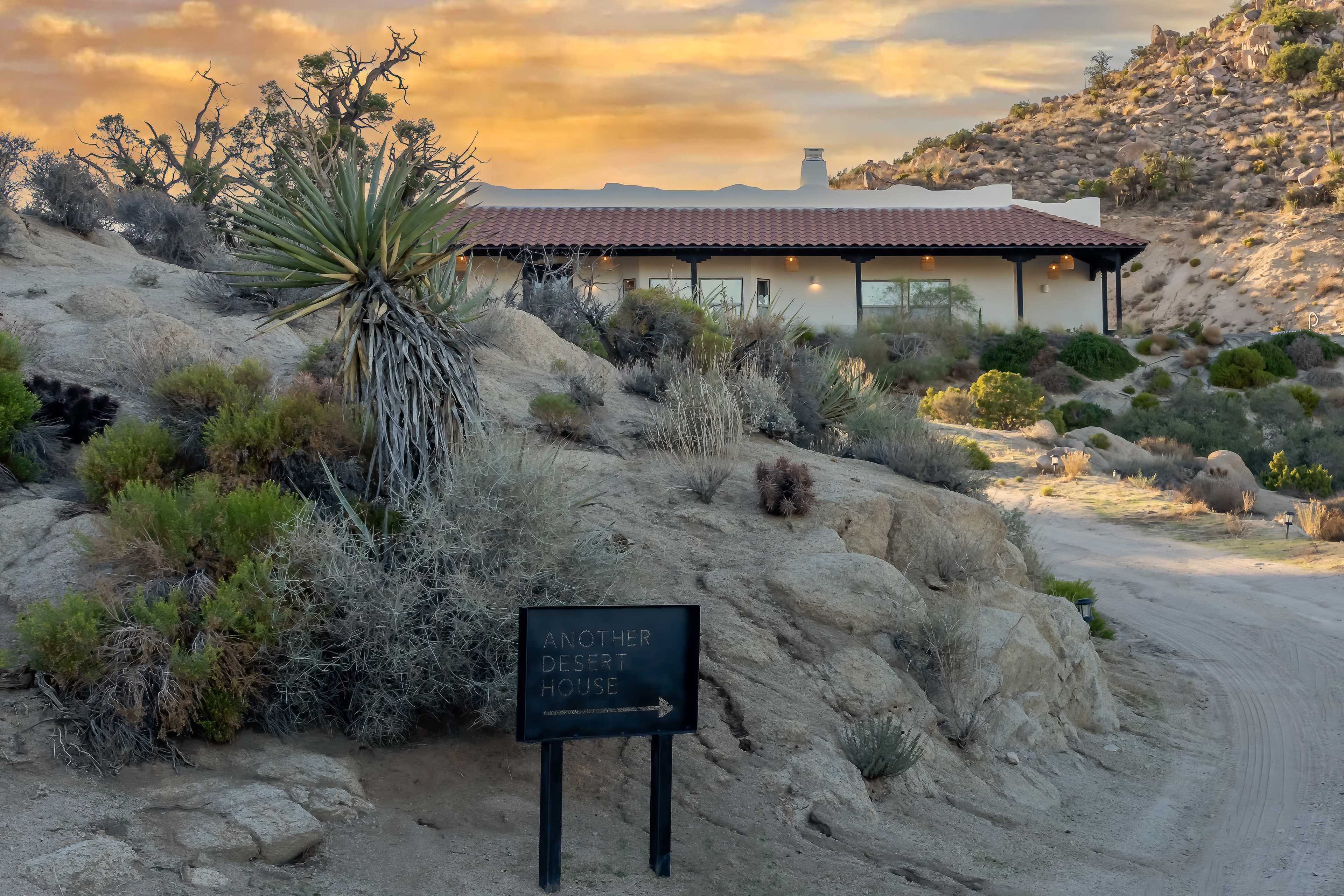 A modern house sits amidst rocky terrain and desert vegetation under a colorful sky.