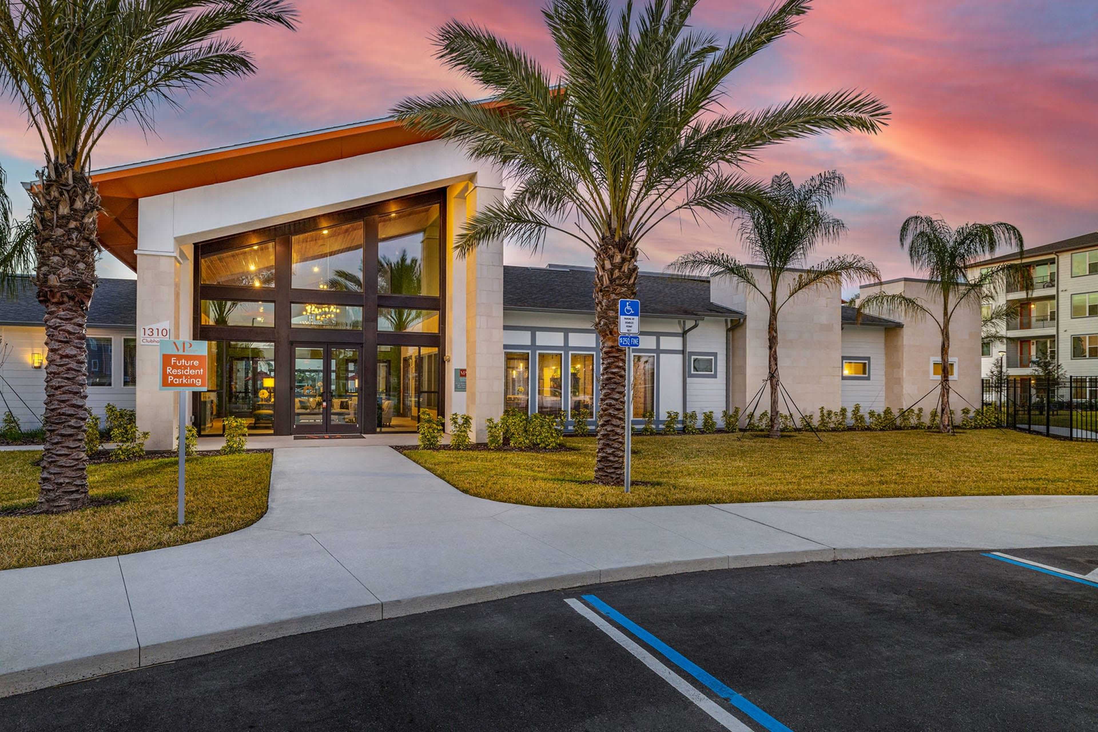 The image shows a modern building entrance with large glass doors, palm trees in front, and a colorful sunset in the background.