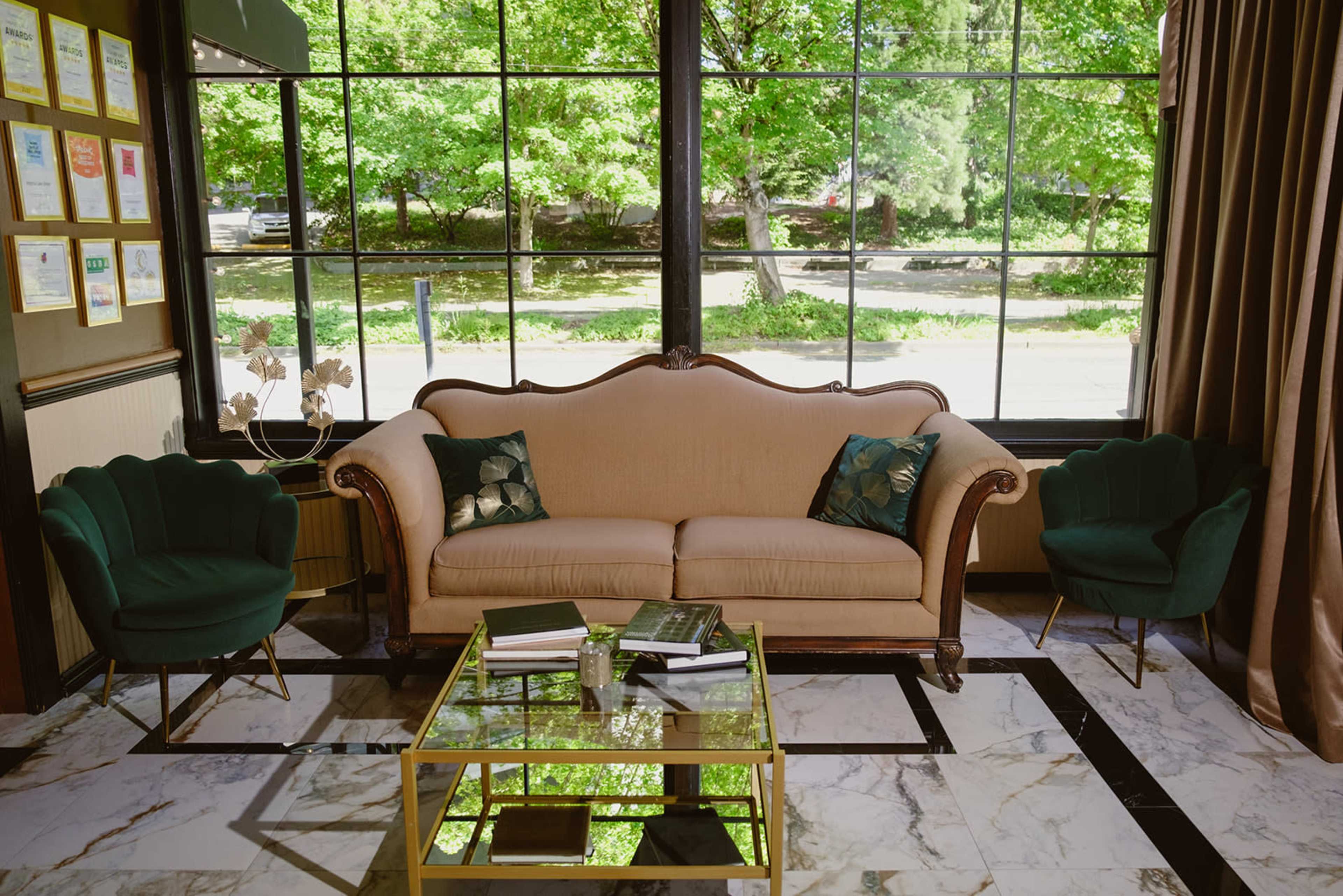 The image shows a living room with a beige sofa, two green armchairs, and a glass coffee table, all set against large windows that offer a view of greenery outside.