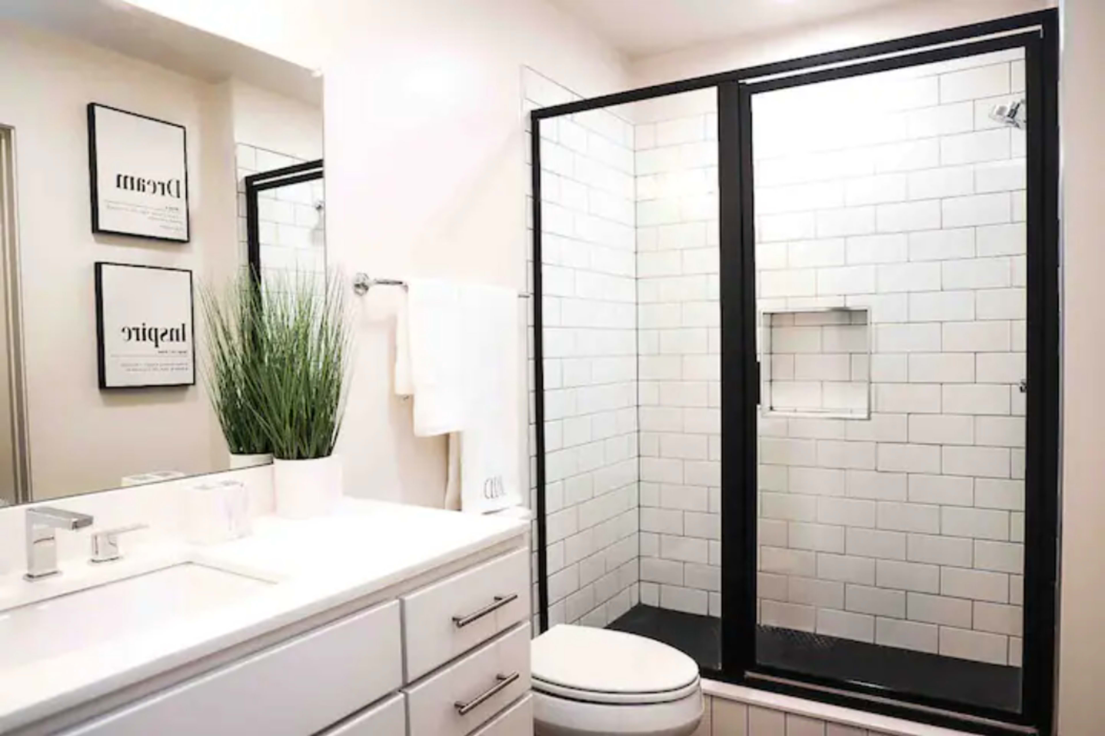 A modern bathroom with a glass shower enclosure, white tiled walls, a white vanity with a sink, and a towel hanging beside a potted plant.