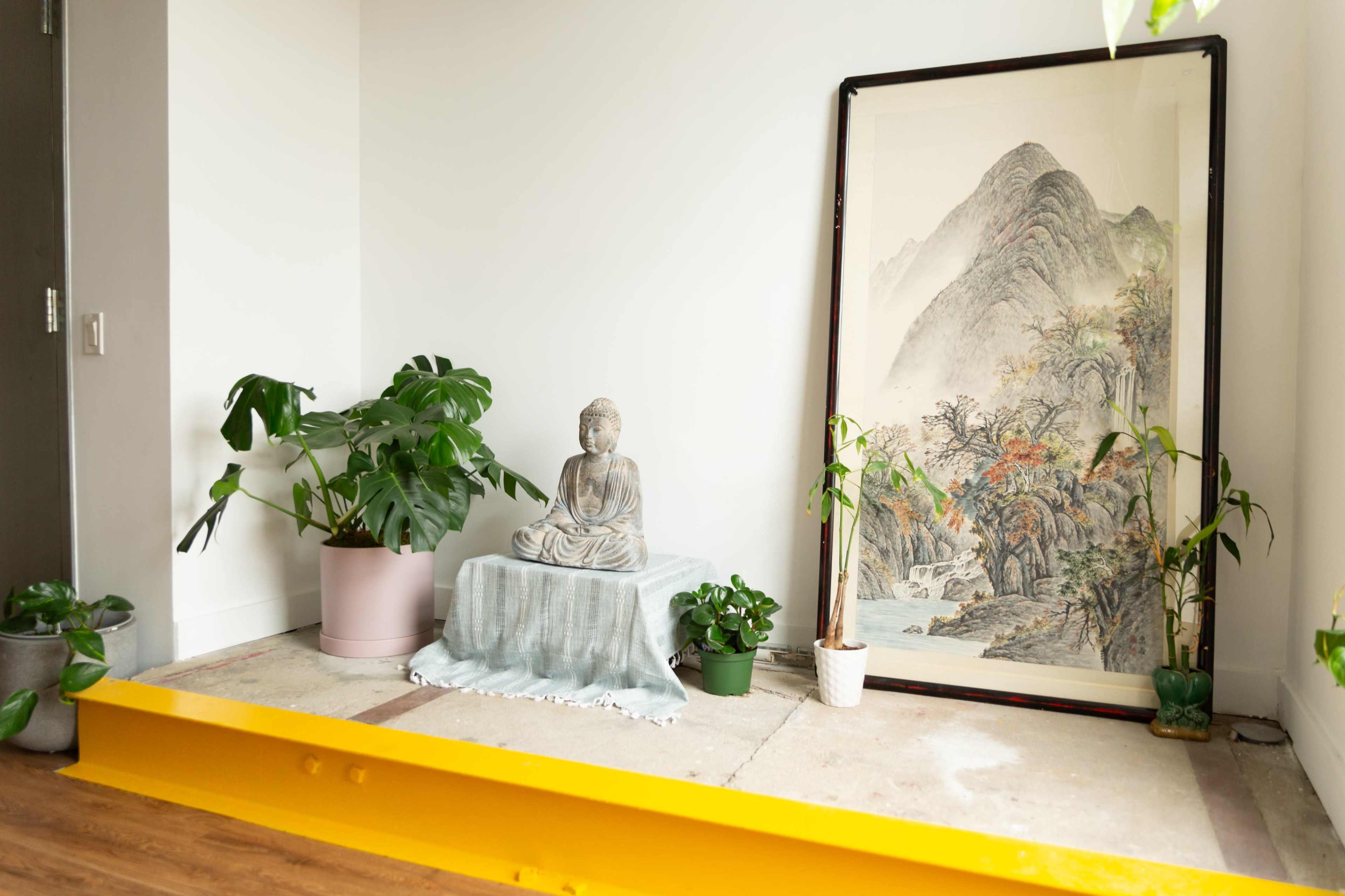 A serene corner of a room features a stone Buddha statue, a framed landscape painting, and several potted plants, arranged on a wooden floor.