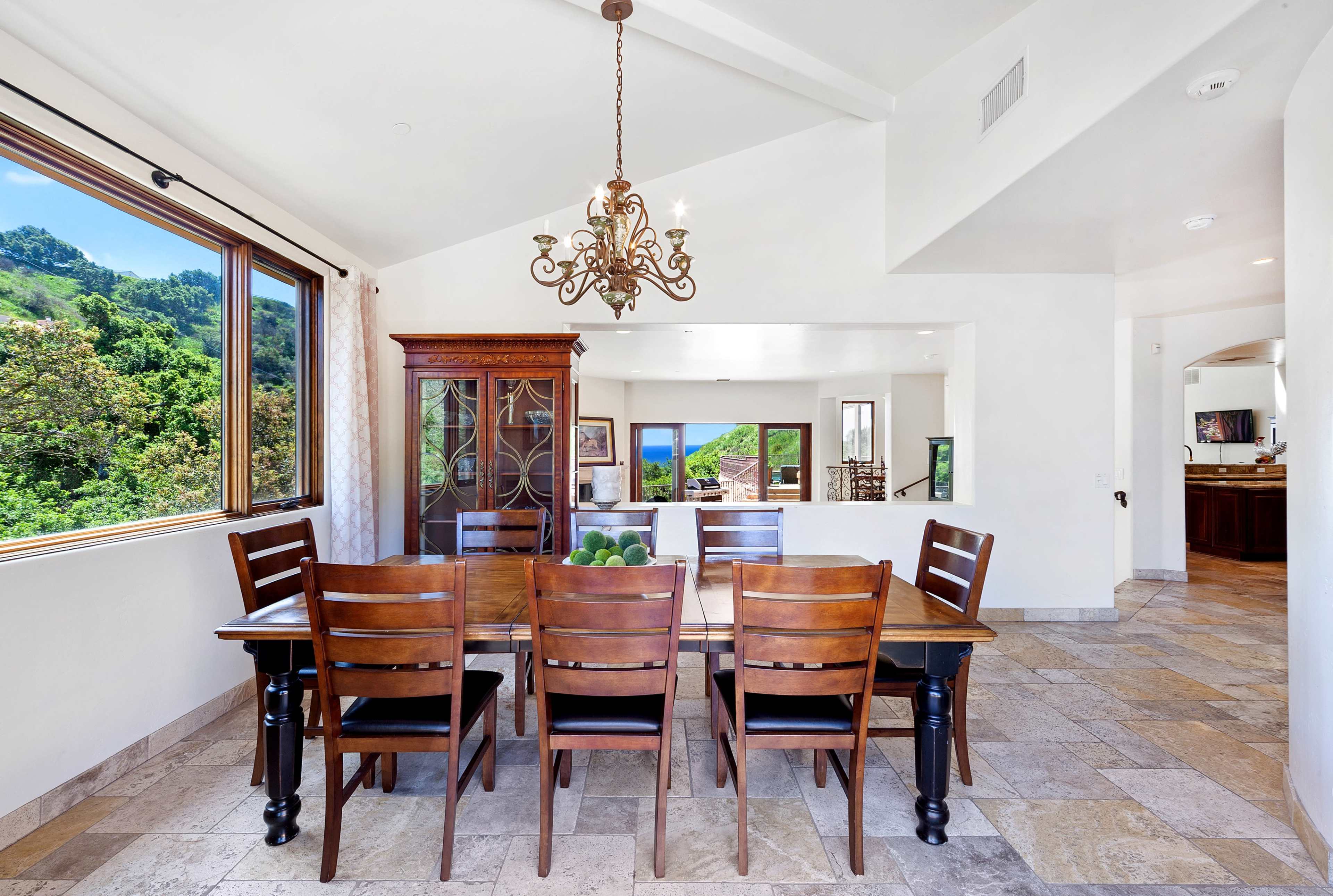The dining area features a wooden table surrounded by chairs, with a chandelier above and large windows showcasing a green hillside outside.