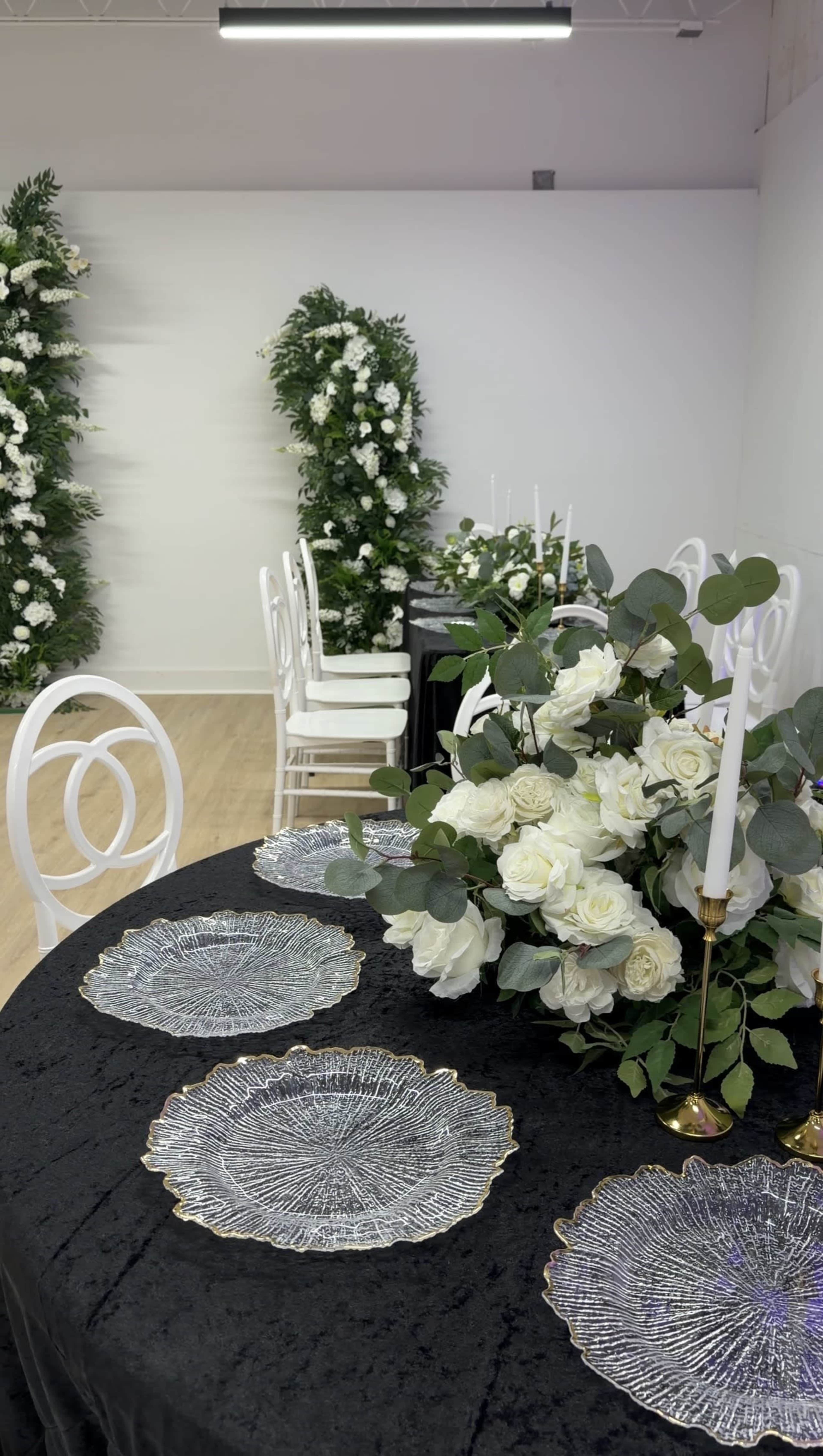 The scene shows a decorated table with elegant white floral arrangements and candles, surrounded by white chairs in a well-lit venue featuring greenery and floral backdrops.