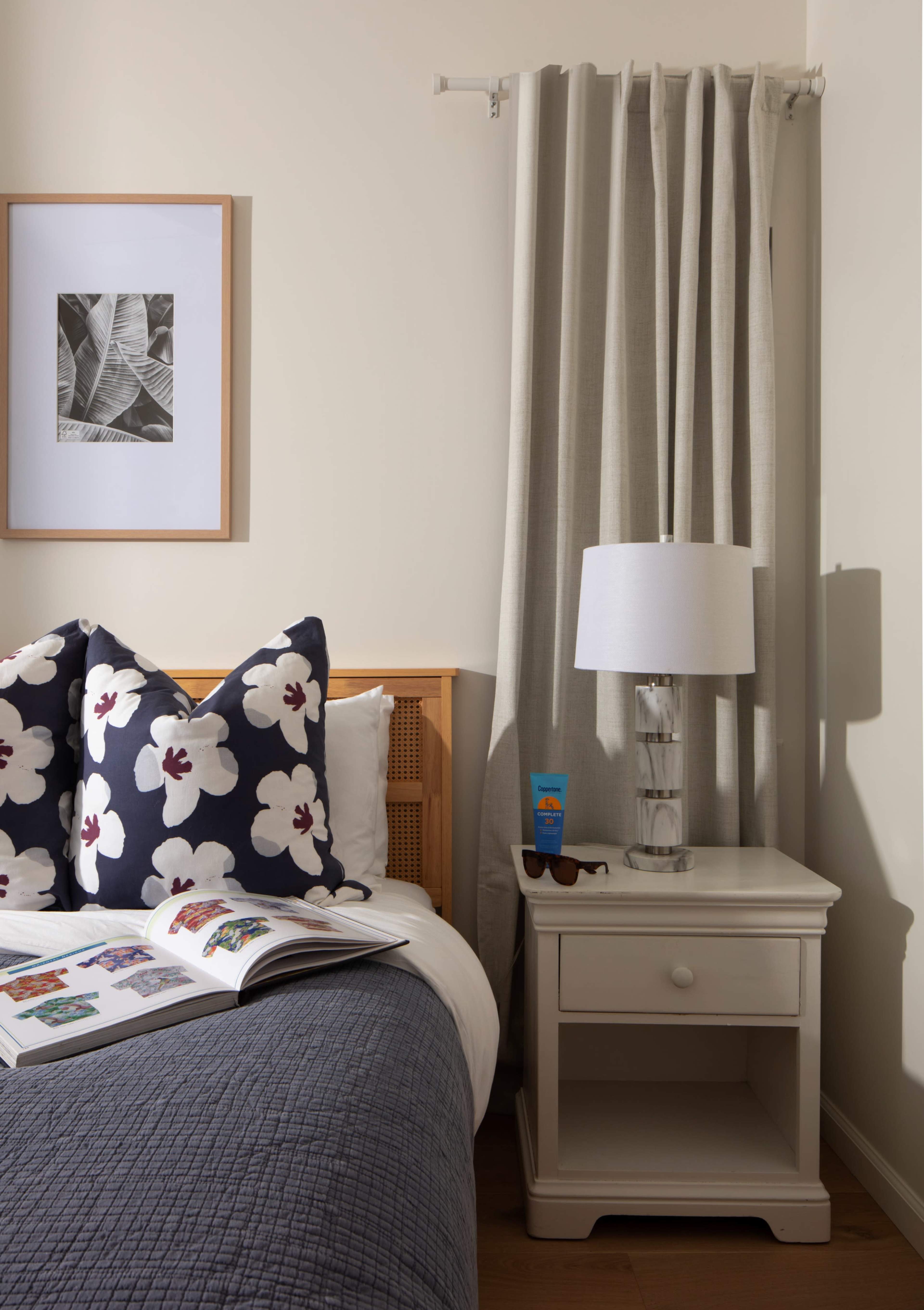 A neatly arranged bedroom features a bed with floral-patterned pillows, a nightstand with a lamp, and a framed artwork on the wall.