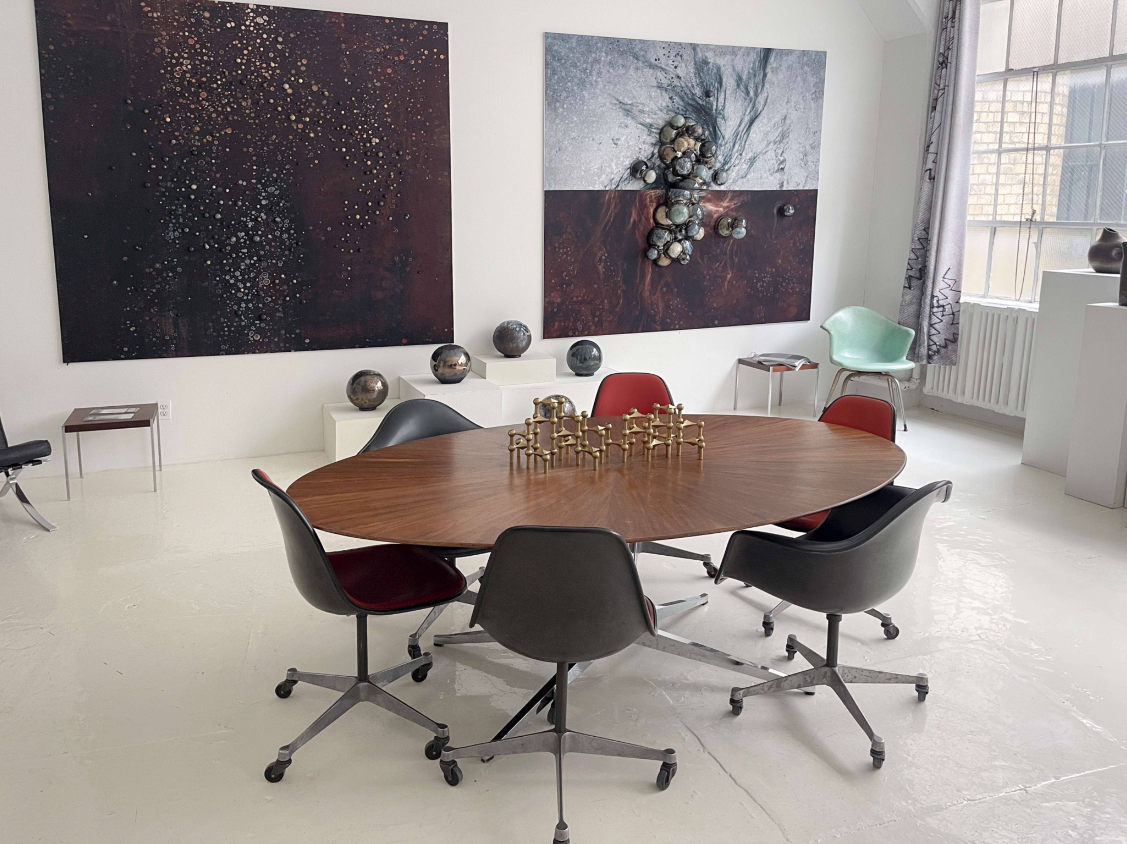 A round wooden table surrounded by six rolling chairs is set in a bright room featuring two large abstract paintings on the walls.