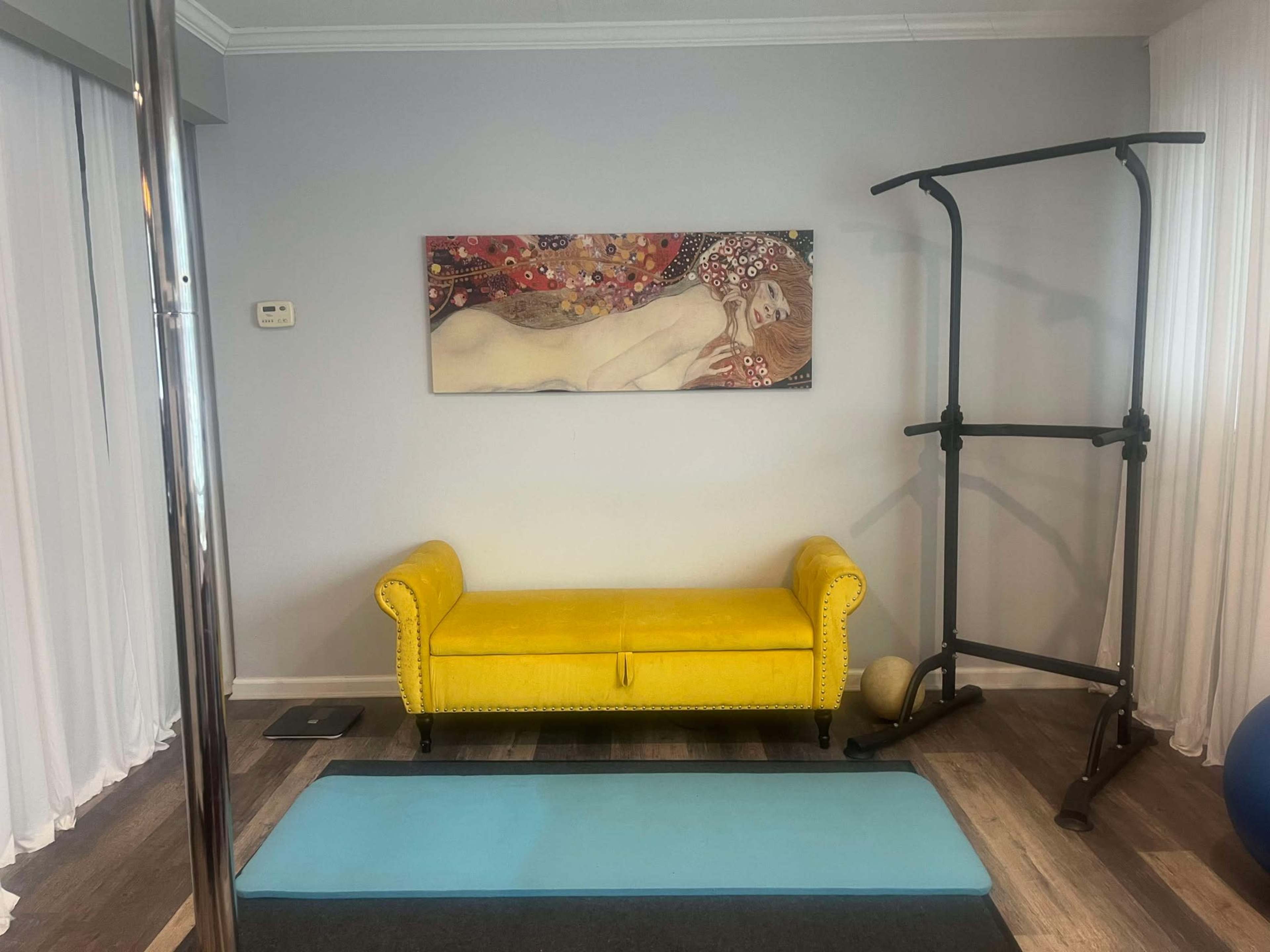 The image shows a yellow couch against a gray wall, a painting above it, a yoga mat on the floor, and a pull-up bar in the corner.