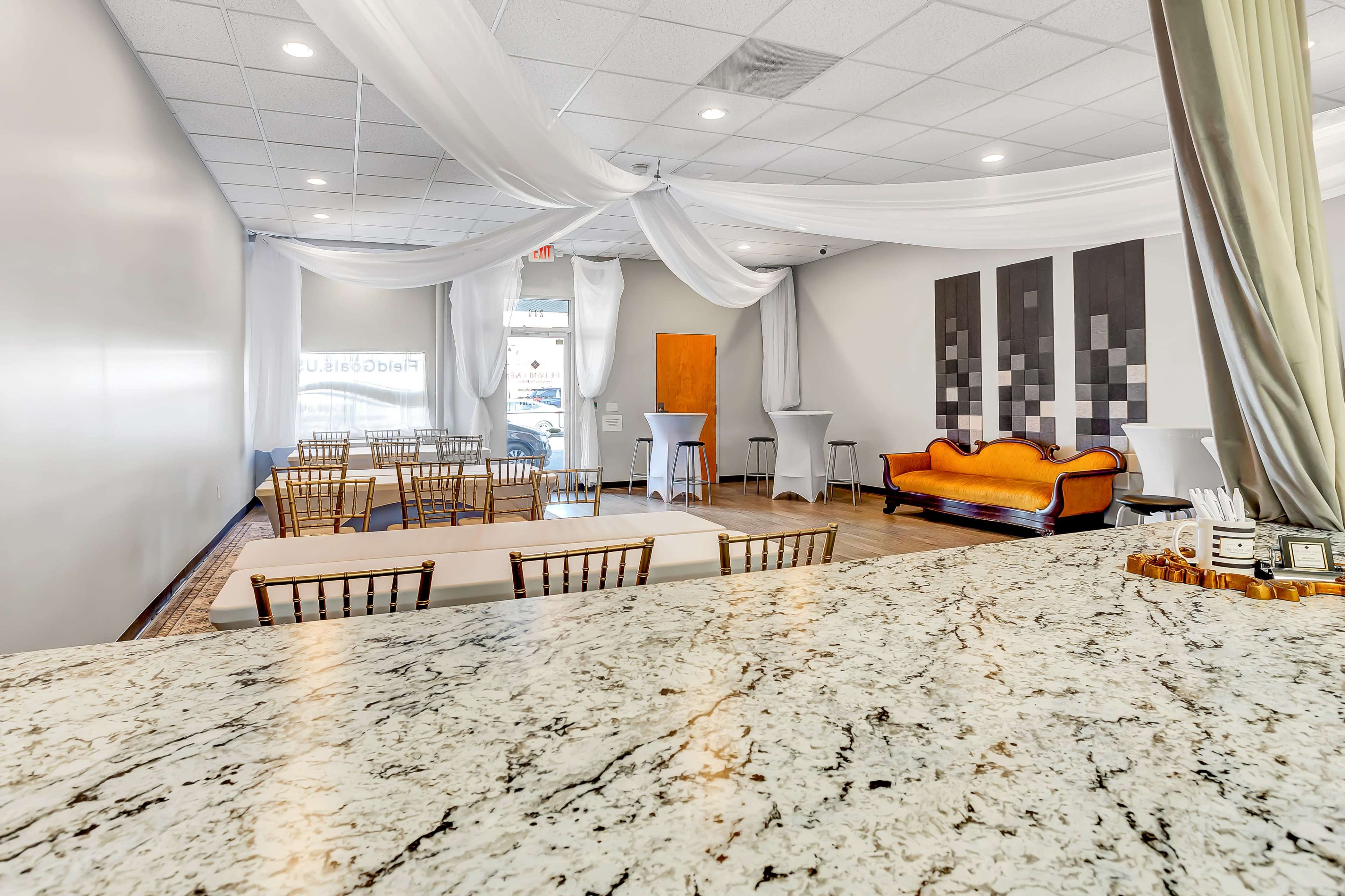 A spacious indoor venue with table and chair setups, draped fabric on the ceiling, and a decorative orange sofa along one wall.