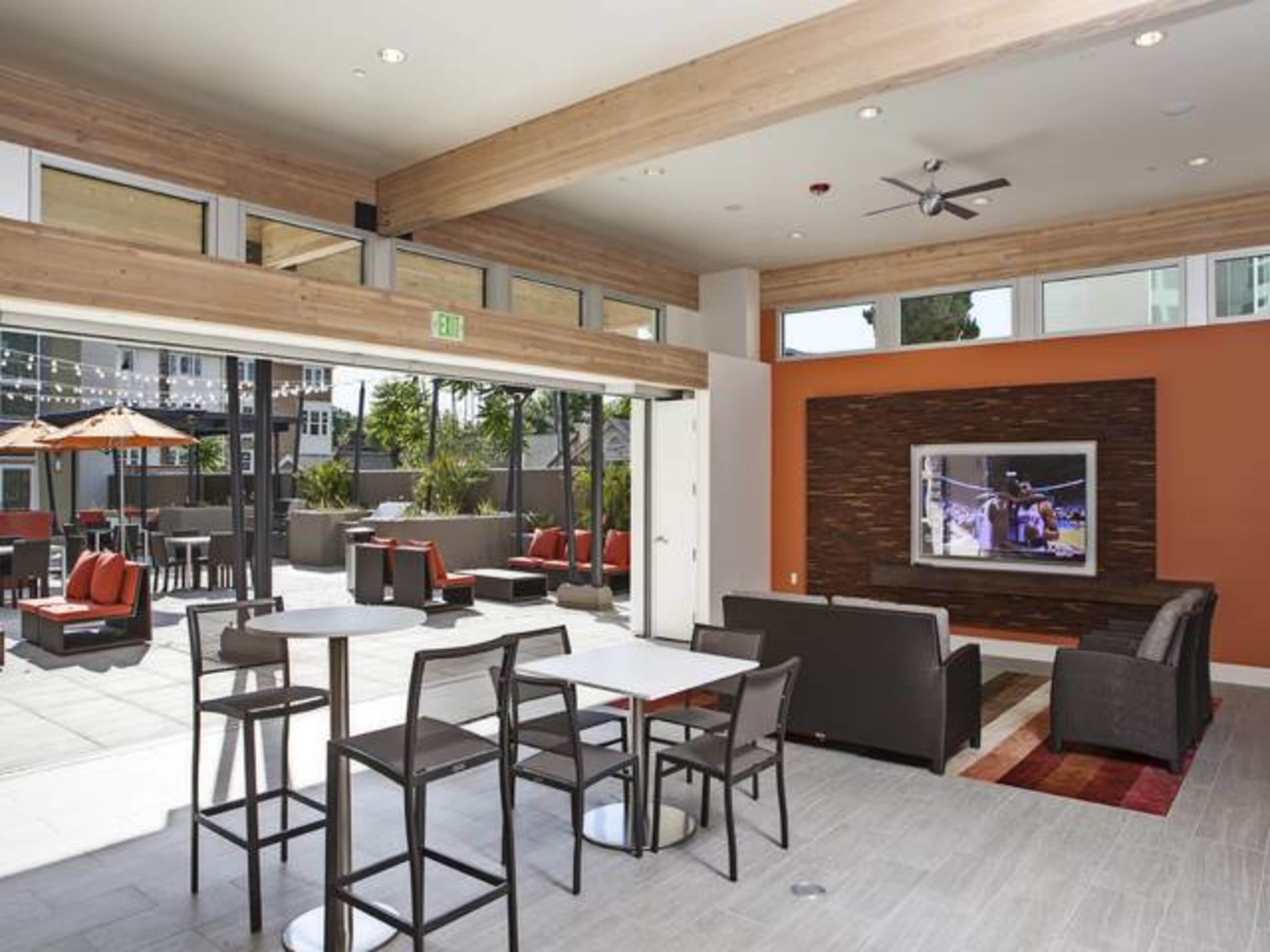 A modern indoor lounge area with a seating arrangement and a large television mounted on the wall, overlooking an outdoor space with patio furniture.