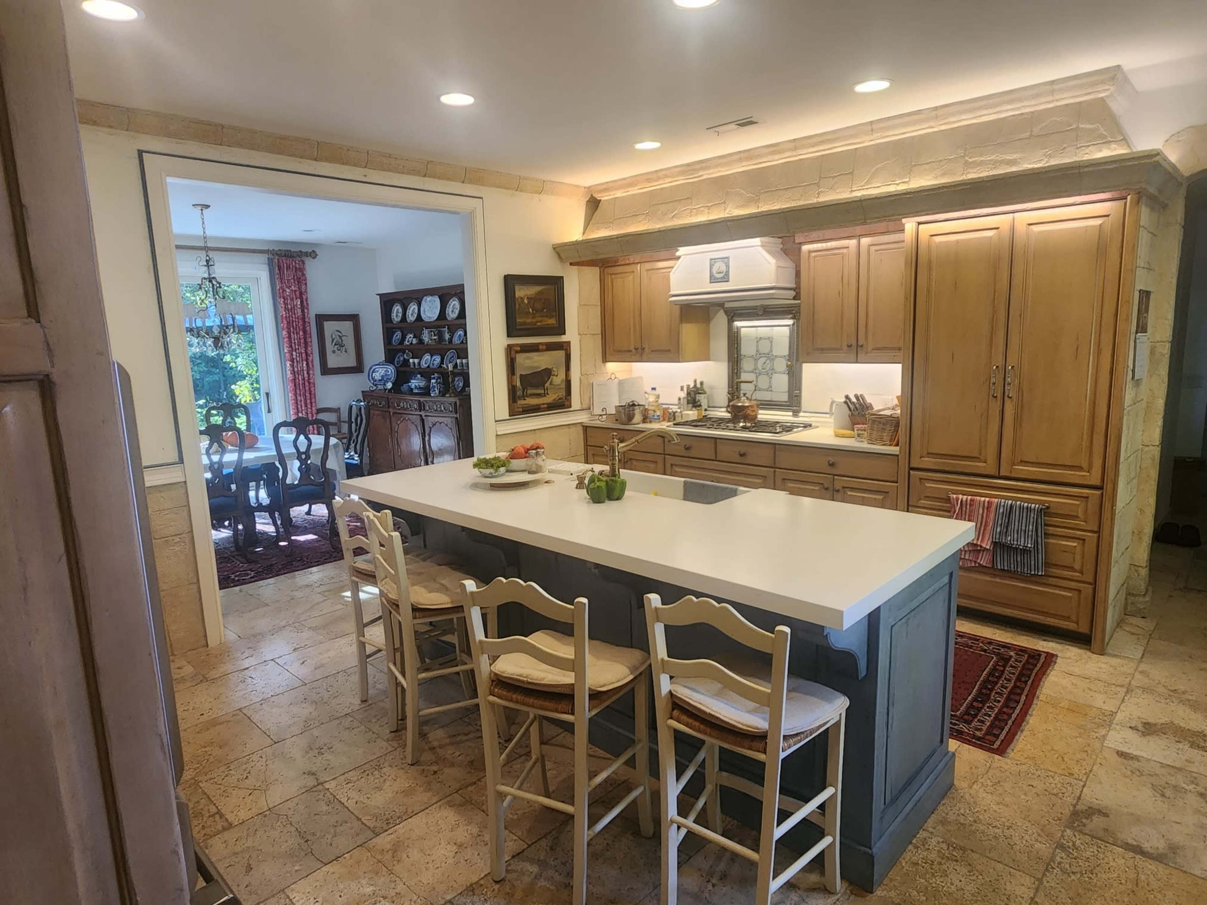 The image shows a spacious kitchen with wooden cabinets, a white island counter with seating, and a view into a dining area furnished with a table and chairs.
