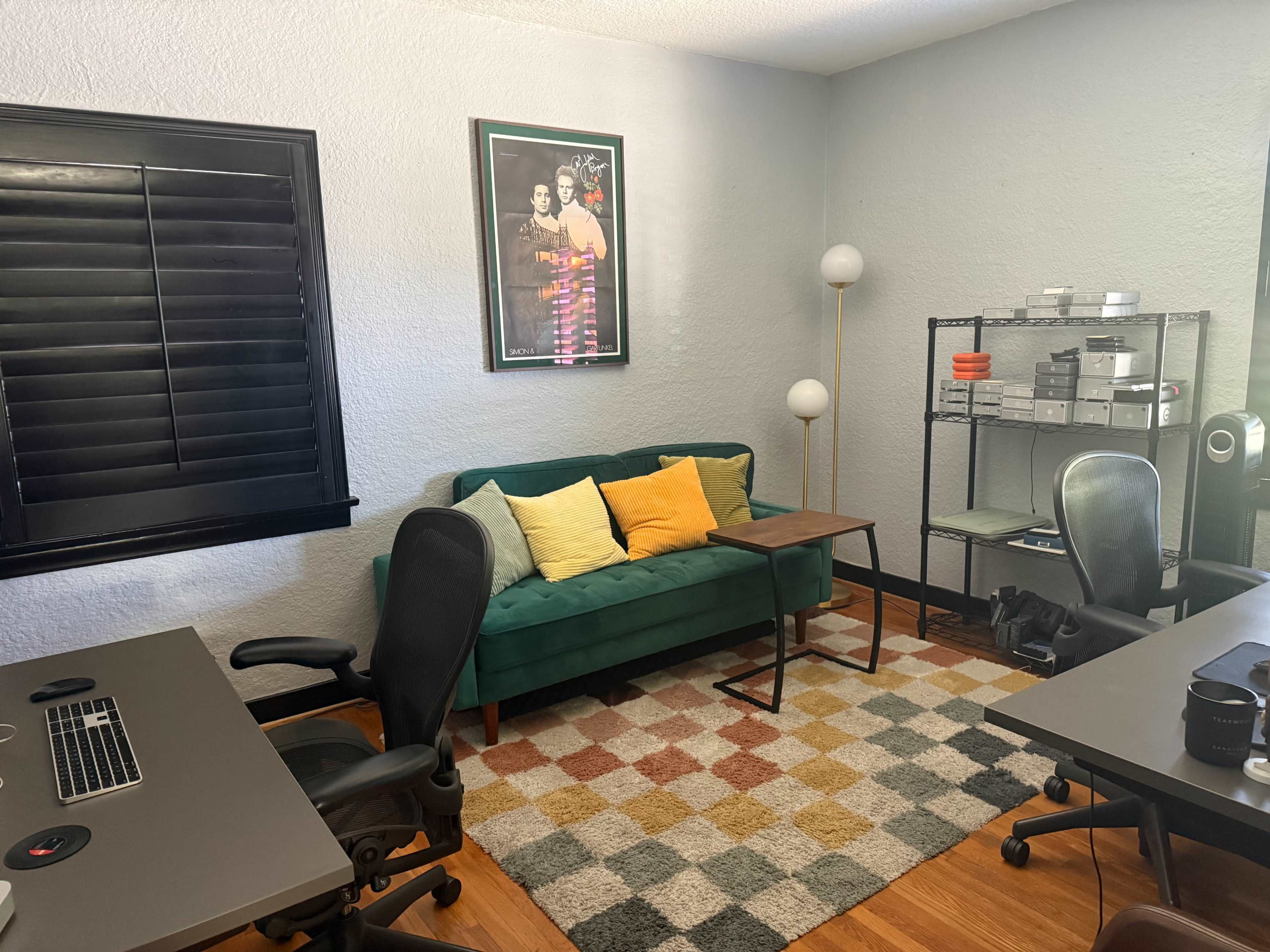 The image shows a modern office space with a green couch, colorful throw pillows, a small side table, and desks with office chairs positioned around a patterned rug.