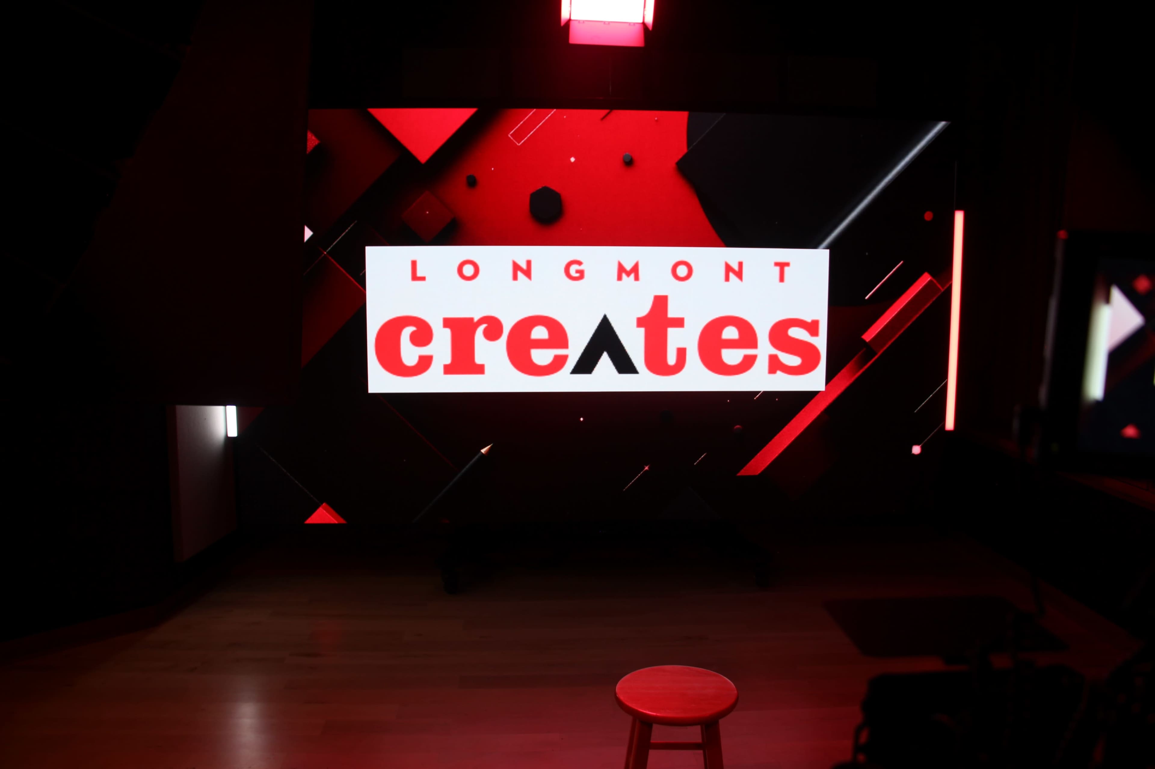 The image features a large screen displaying the text "LONGMONT creates" with a geometric design in red, black, and white, alongside a wooden stool in the foreground.