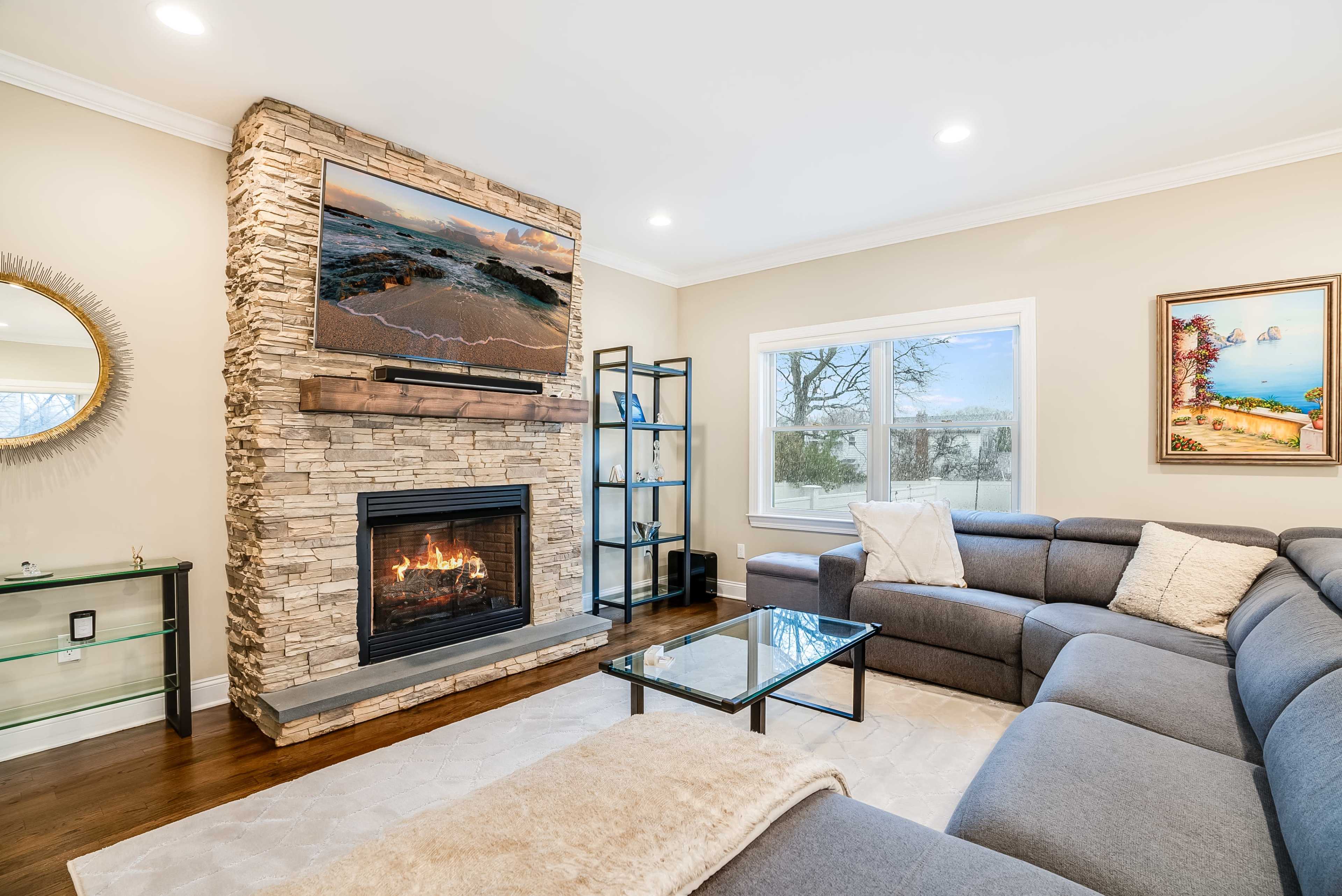 A living room with a stone fireplace, a mounted television above the mantel, and a gray sectional sofa facing a glass coffee table.