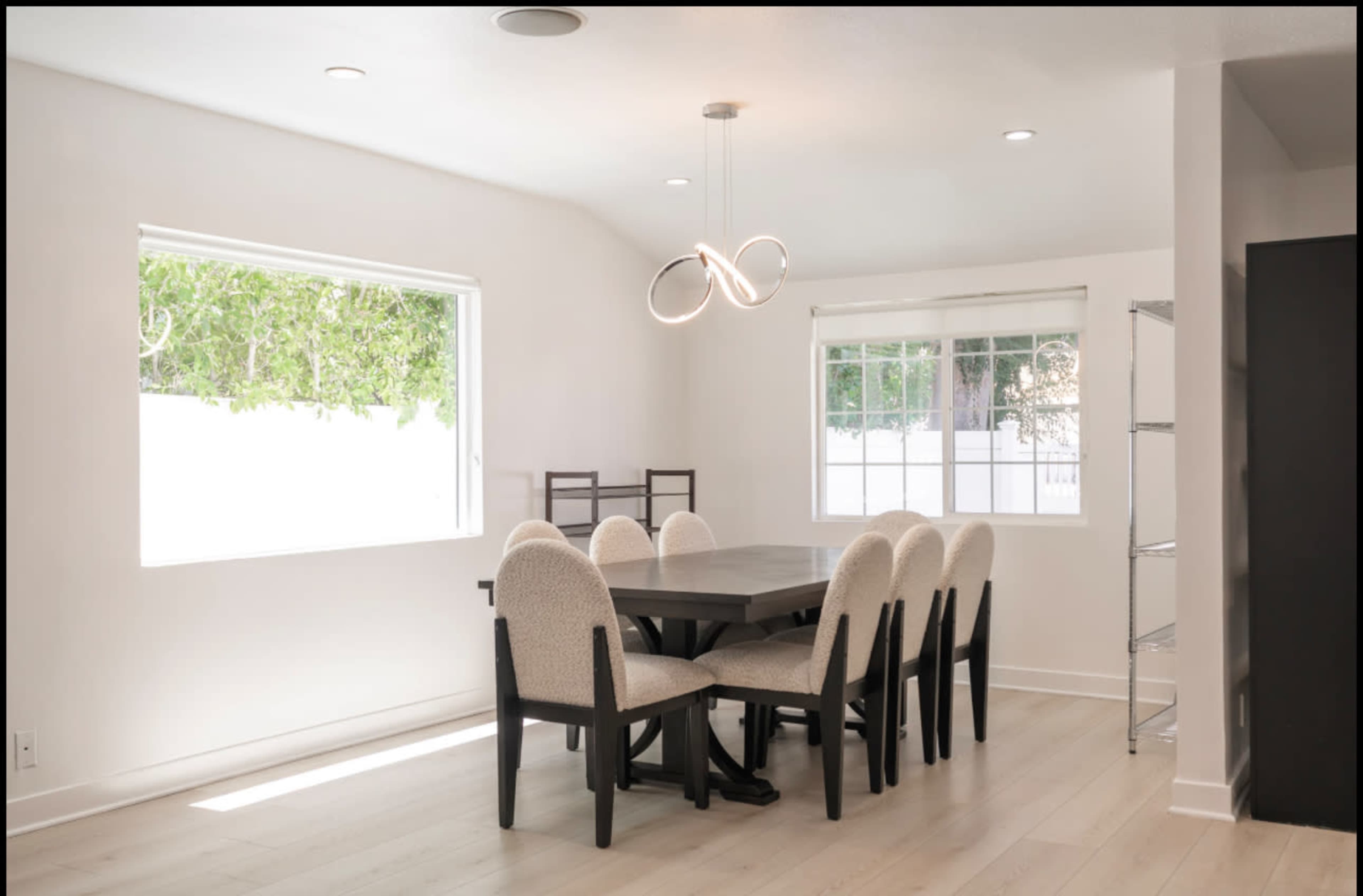 A dining area features a large wooden table surrounded by upholstered chairs, with a modern light fixture above and windows letting in natural light.