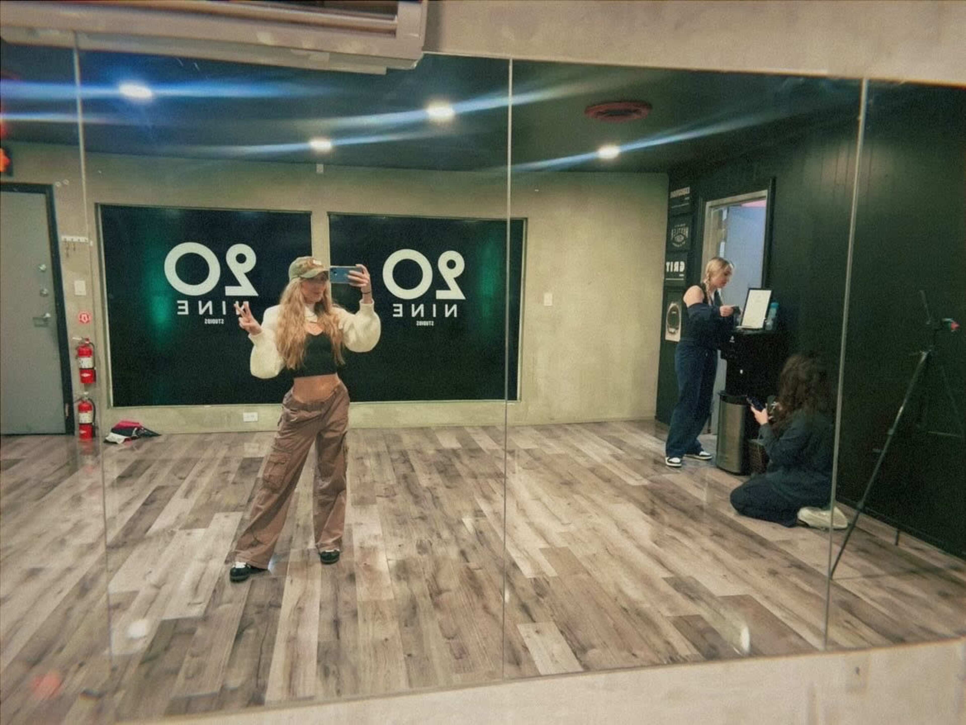 Three individuals in a dance studio, with one person posing in front of a mirror while the others are interacting with a sound system in the background.