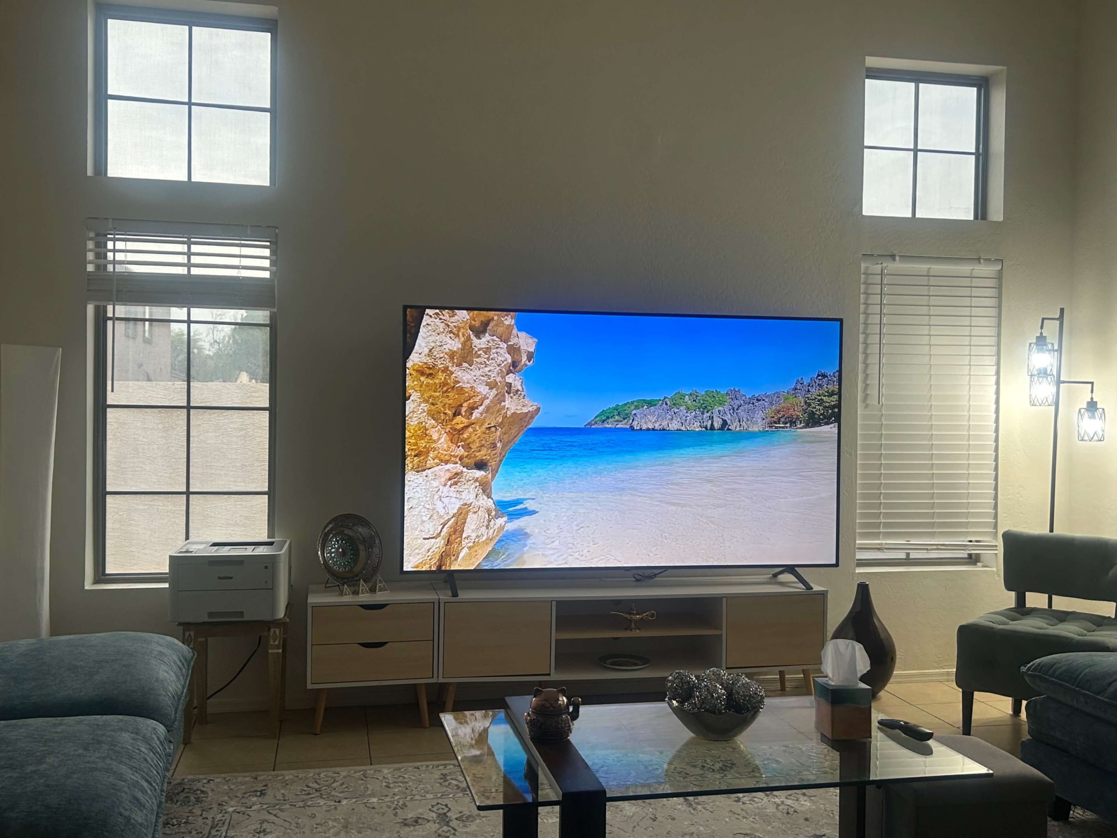 A large television displaying a beach scene is positioned in a living room with windows allowing natural light to enter.