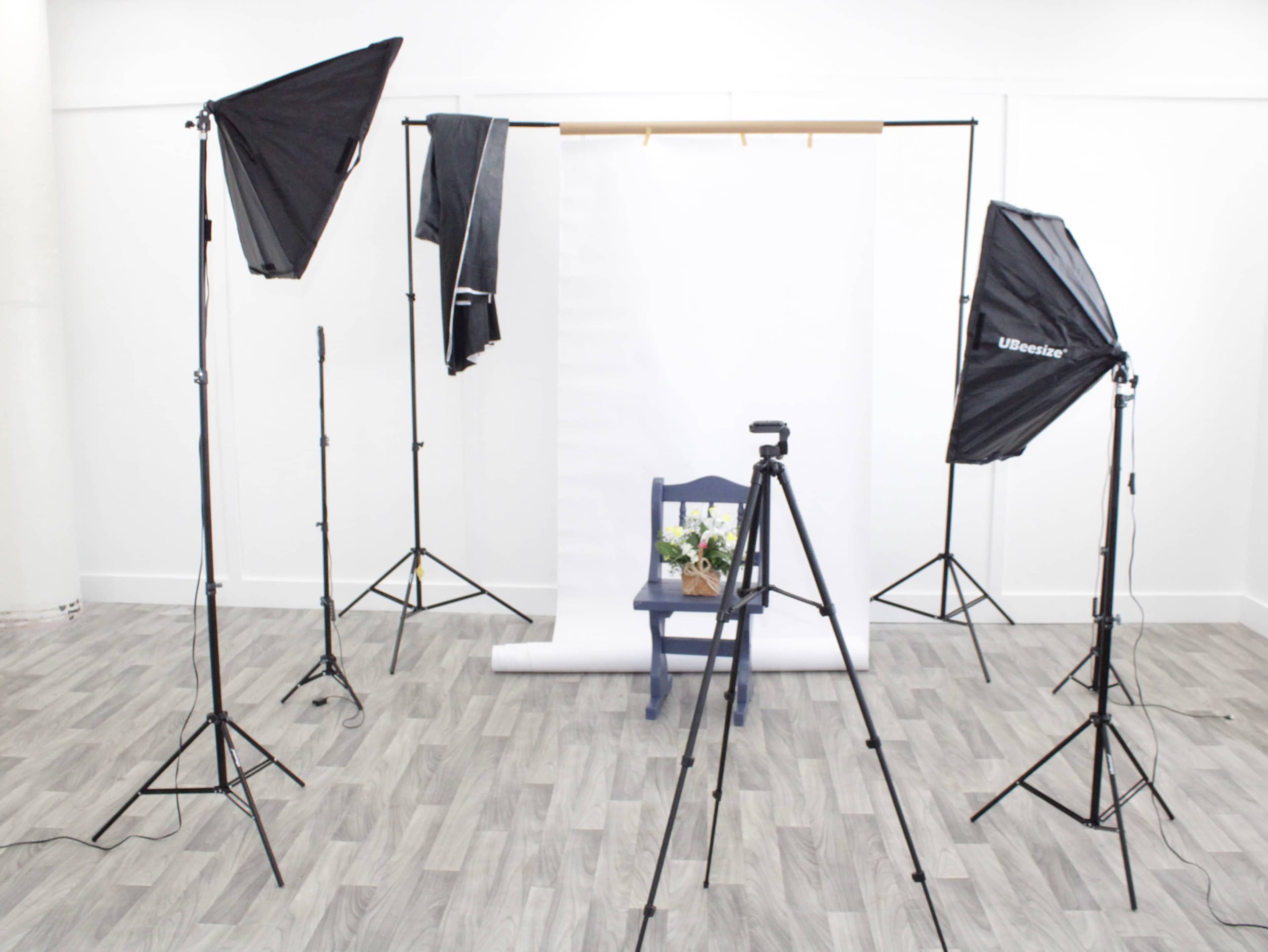 The image shows a photography setup with two softboxes, a camera on a tripod, and a blue chair adorned with flowers positioned in front of a plain white backdrop.