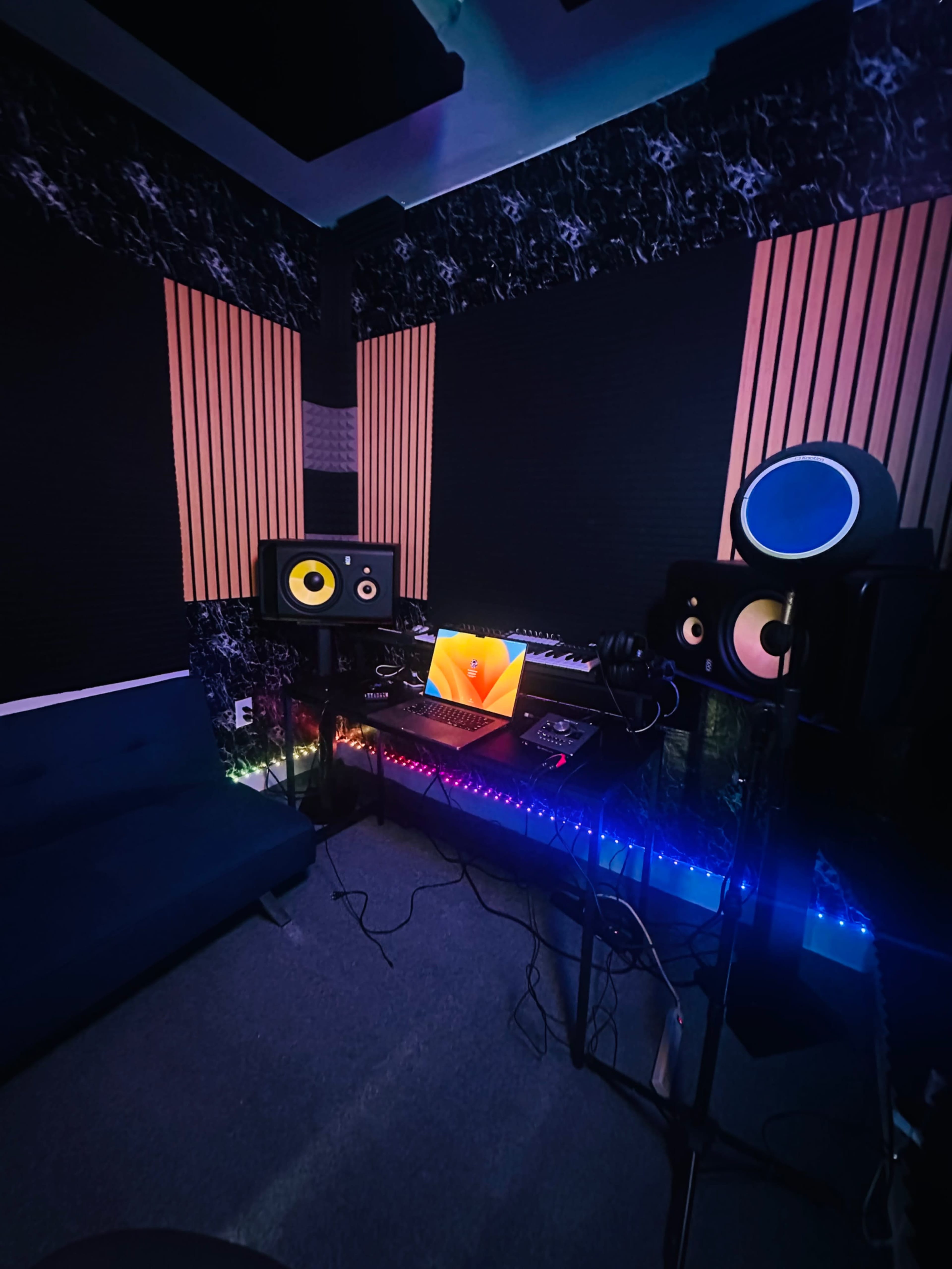 A music production studio features a computer setup with speakers, a keyboard, and colorful LED lighting.