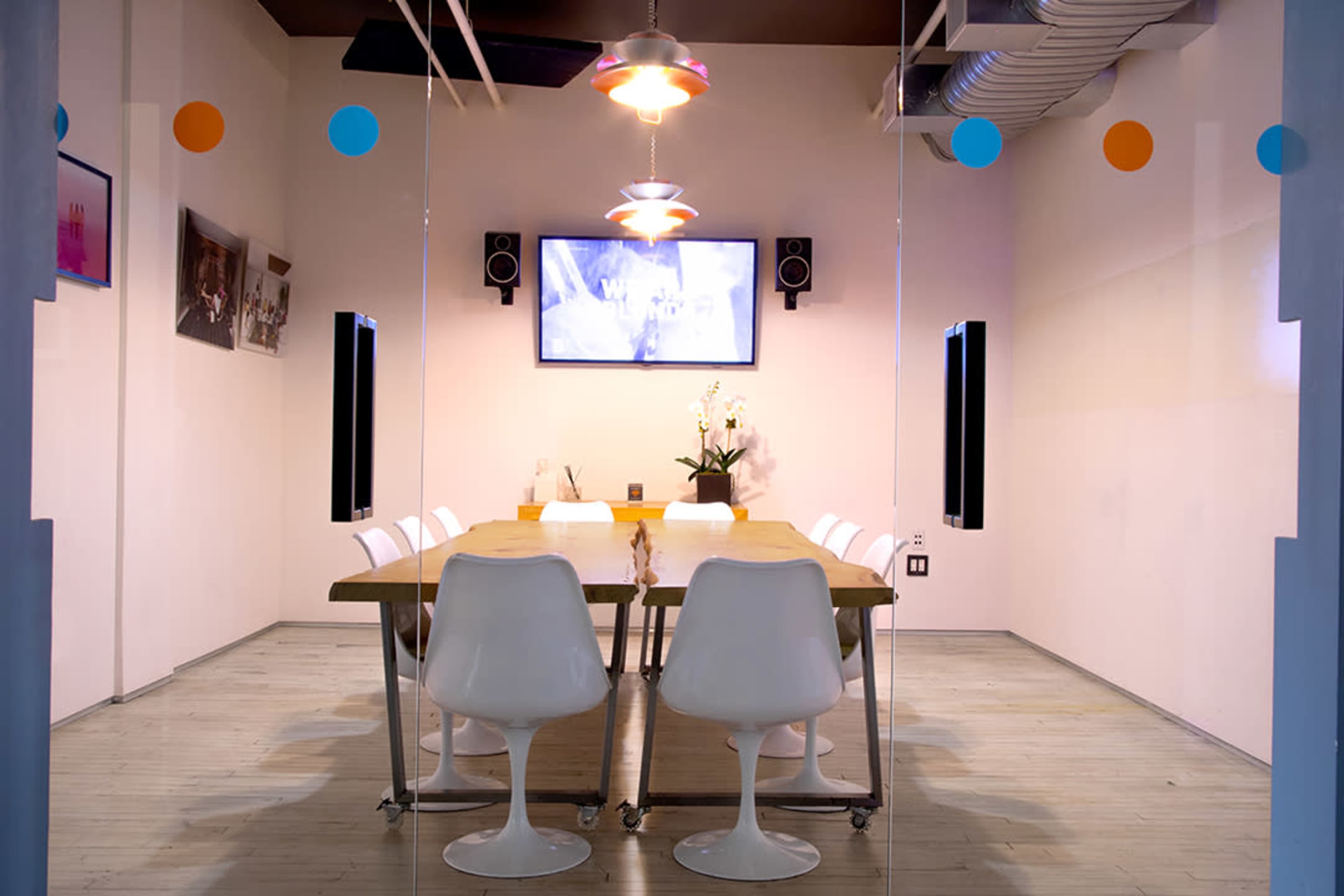 A conference room features a large wooden table surrounded by white chairs, with a wall-mounted screen and audio speakers in the background.