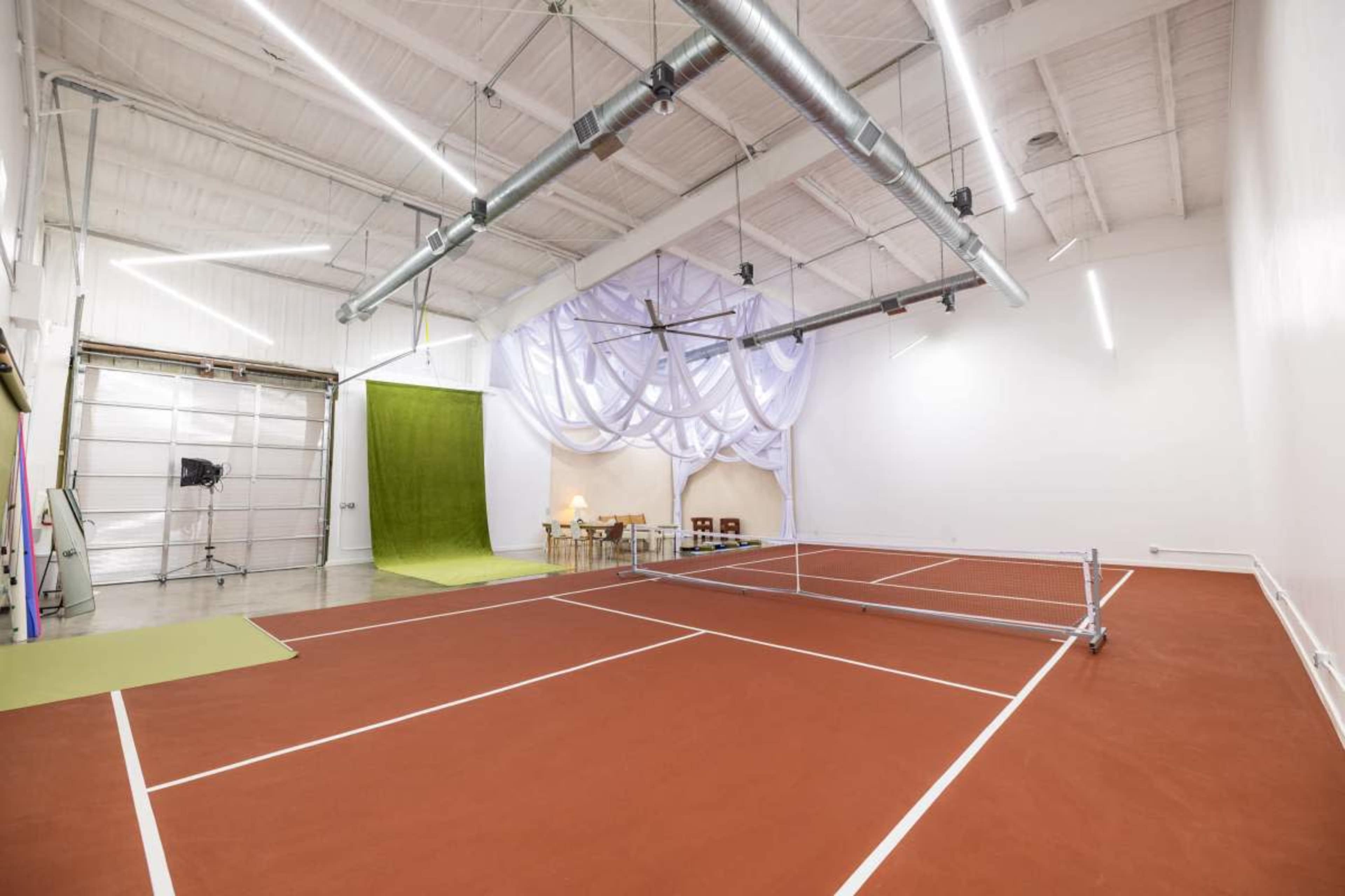 The image shows an indoor space featuring a tennis court with red flooring, a green backdrop, and studio lighting.