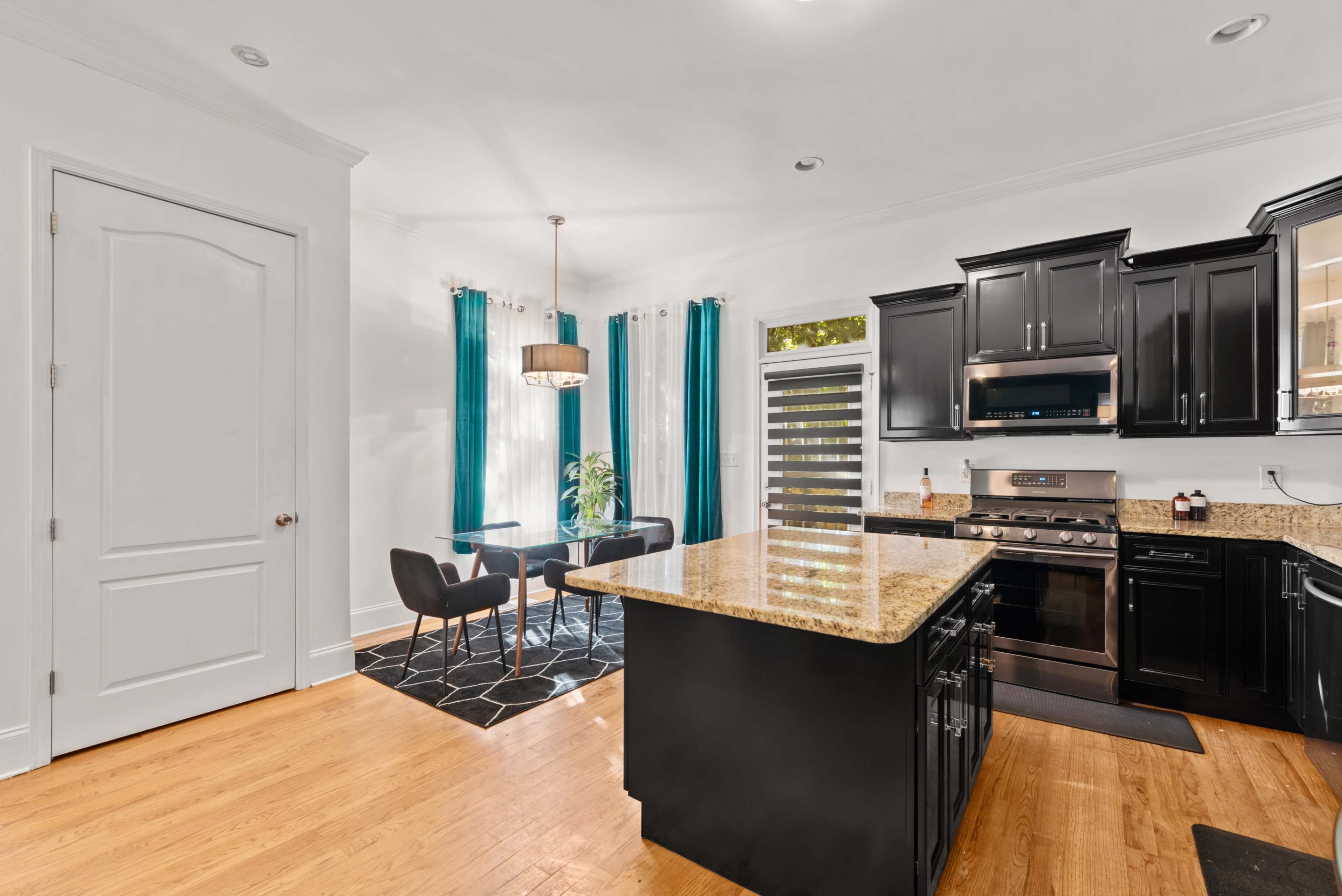 A modern kitchen with black cabinets, a granite island, and a dining area featuring a round table and chairs, illuminated by natural light through large windows.