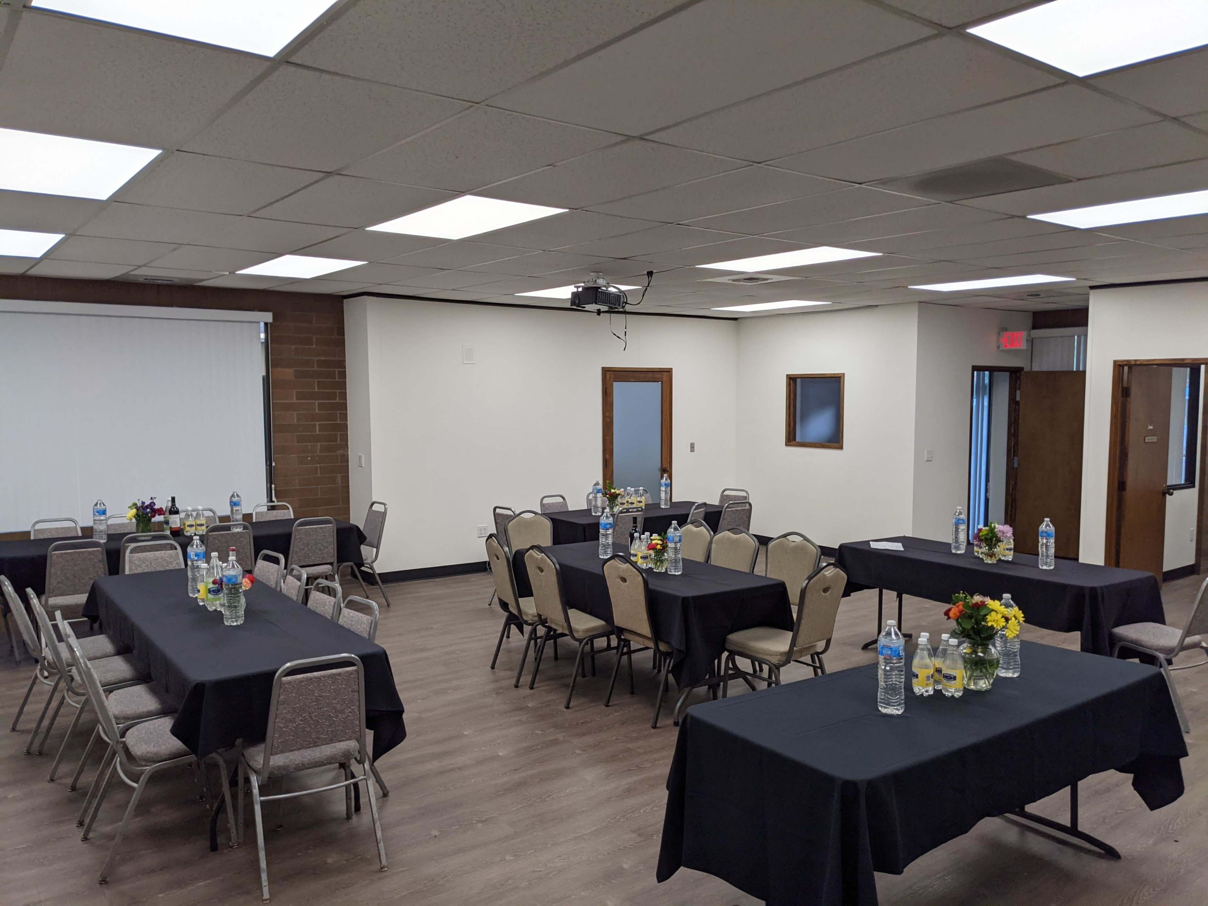 A large meeting room features several tables covered with black cloths, each decorated with floral centerpieces and bottles of water, arranged in a grid layout.
