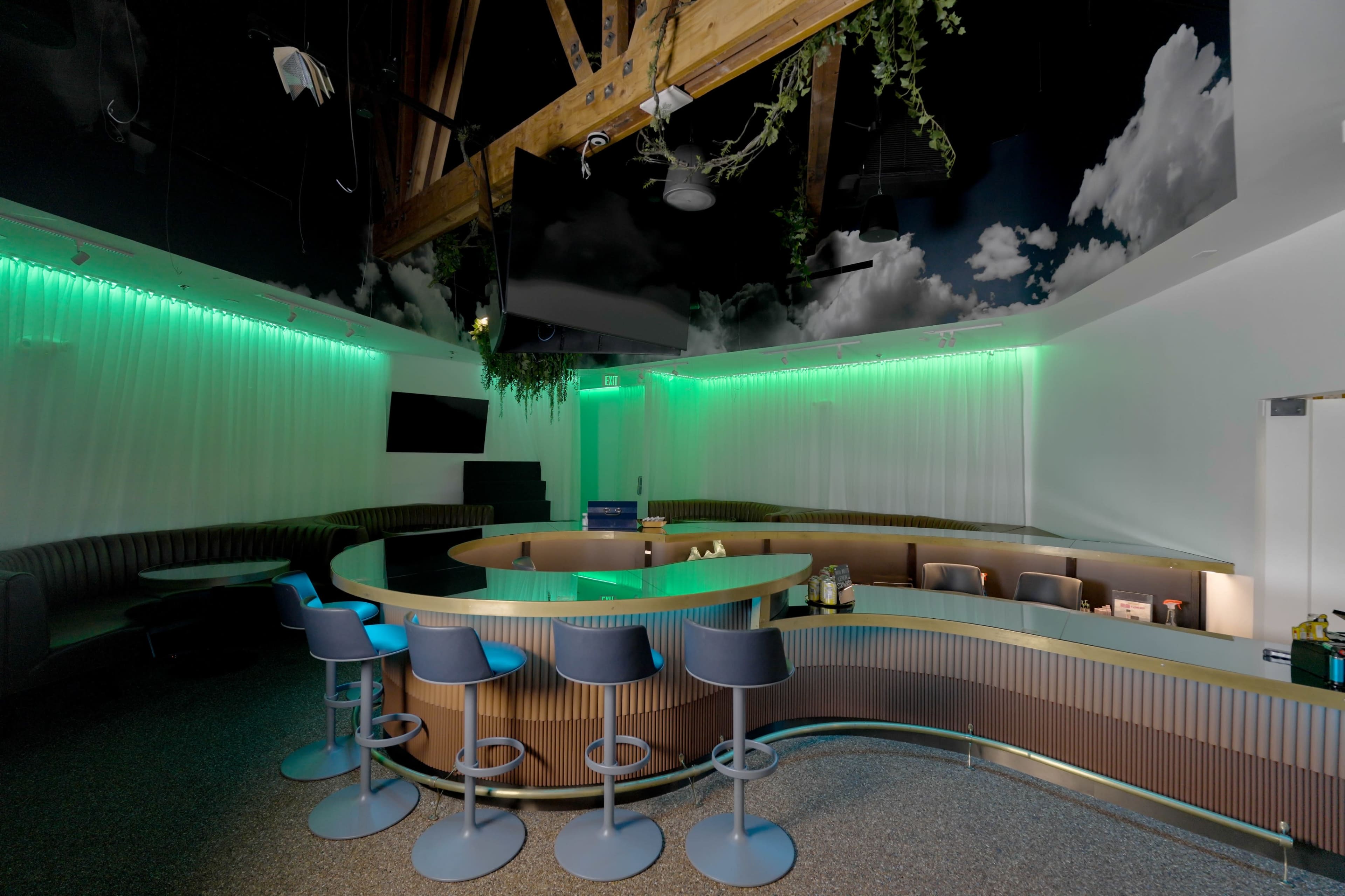The image shows a modern bar area with a curved counter and illuminated green lighting, complemented by a ceiling featuring a cloud design.