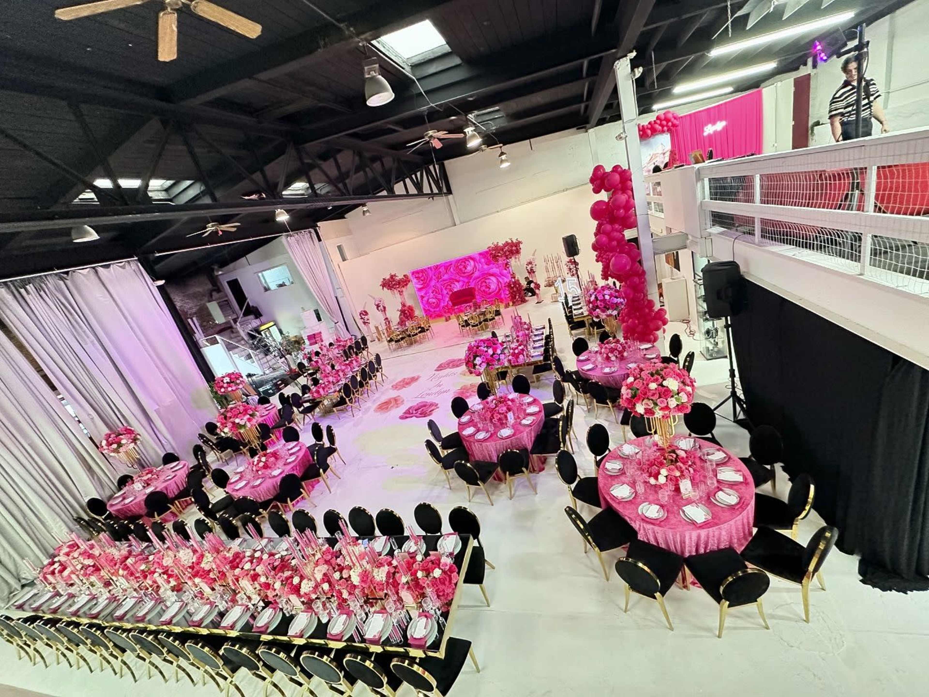 The image shows a large event space decorated in pink, featuring round tables with floral centerpieces and a staged backdrop.
