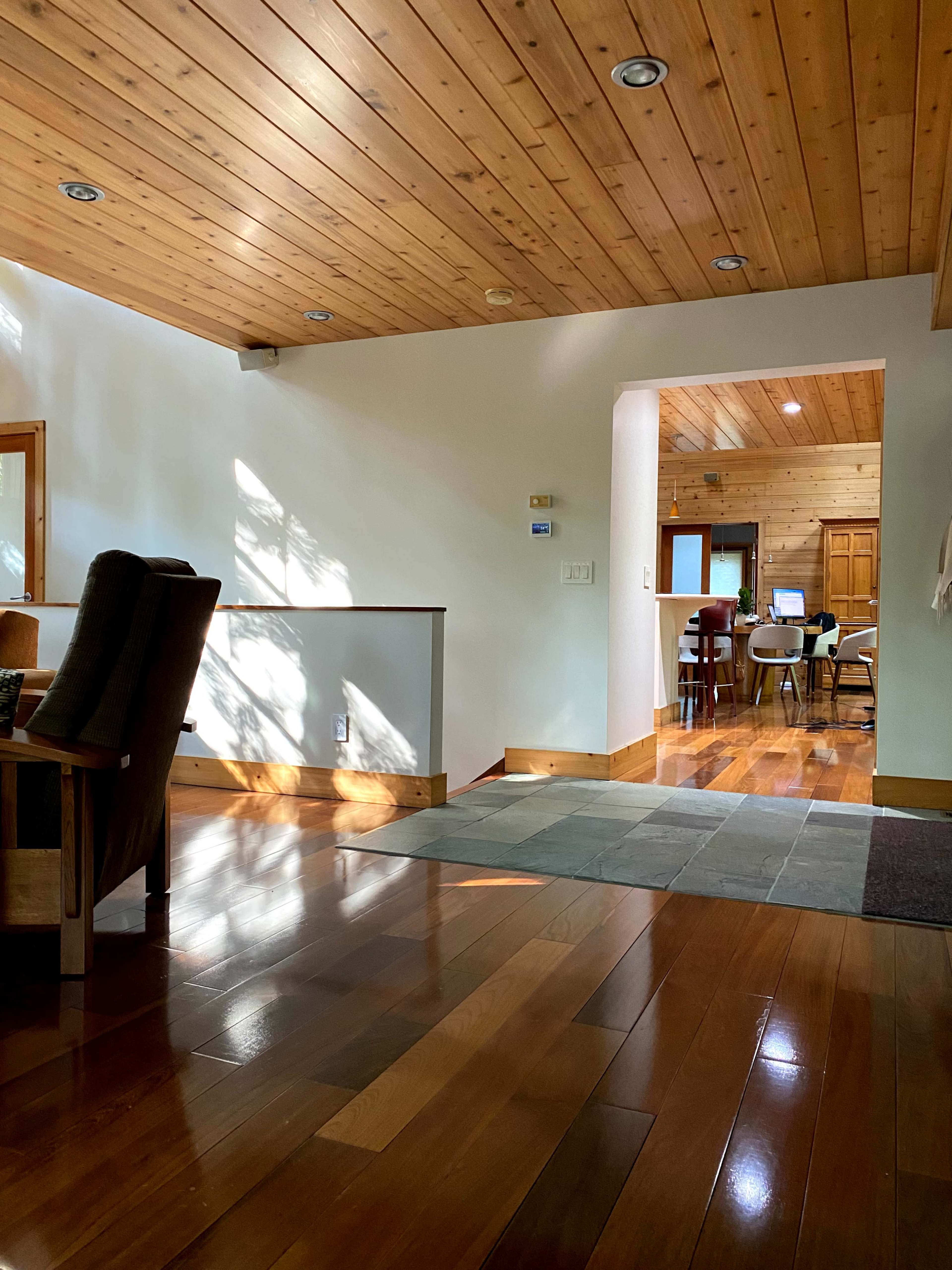 The image shows a well-lit interior space with wooden ceiling beams, a tiled floor, and a view into an adjoining room with wooden furniture.