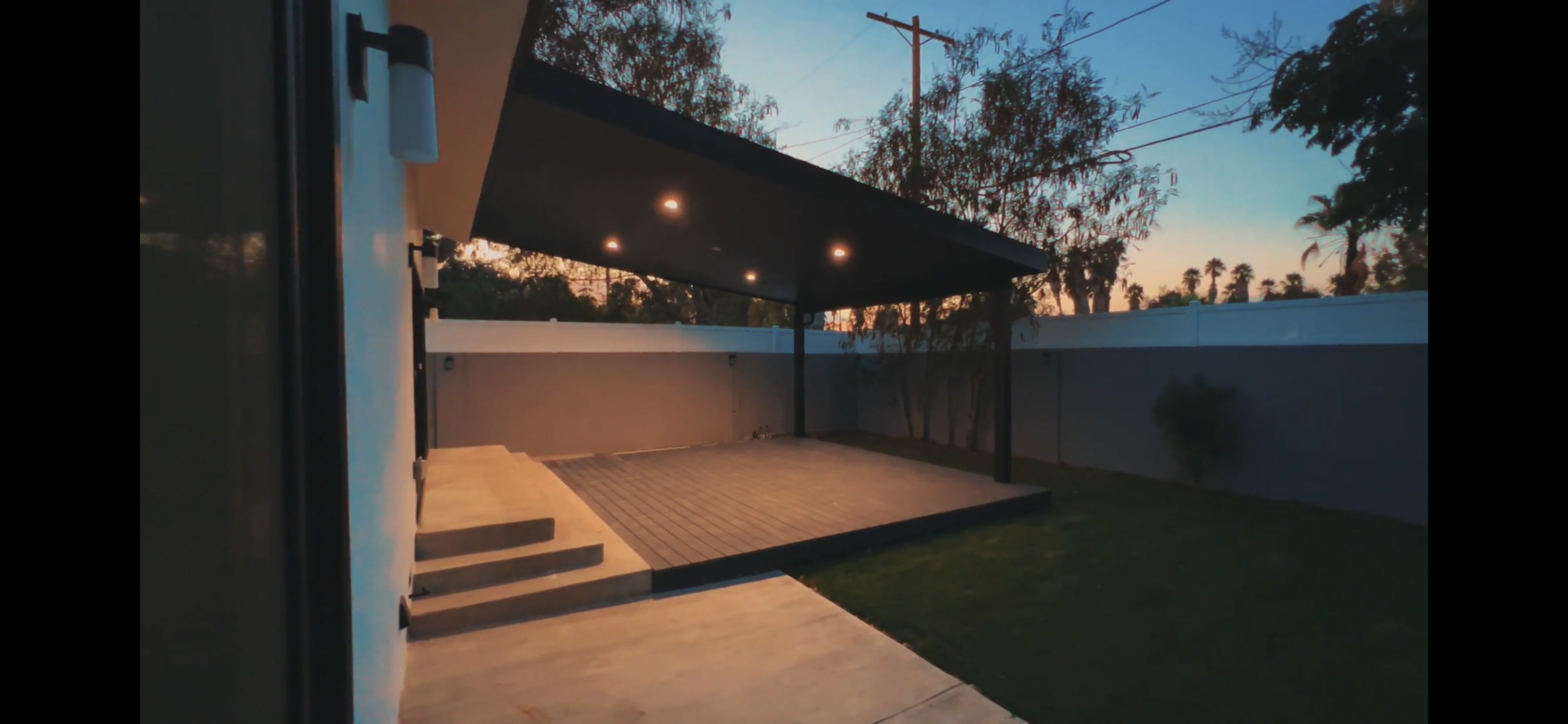 A modern outdoor patio area with a flat roof, wooden decking, and a grassy lawn, during dusk.