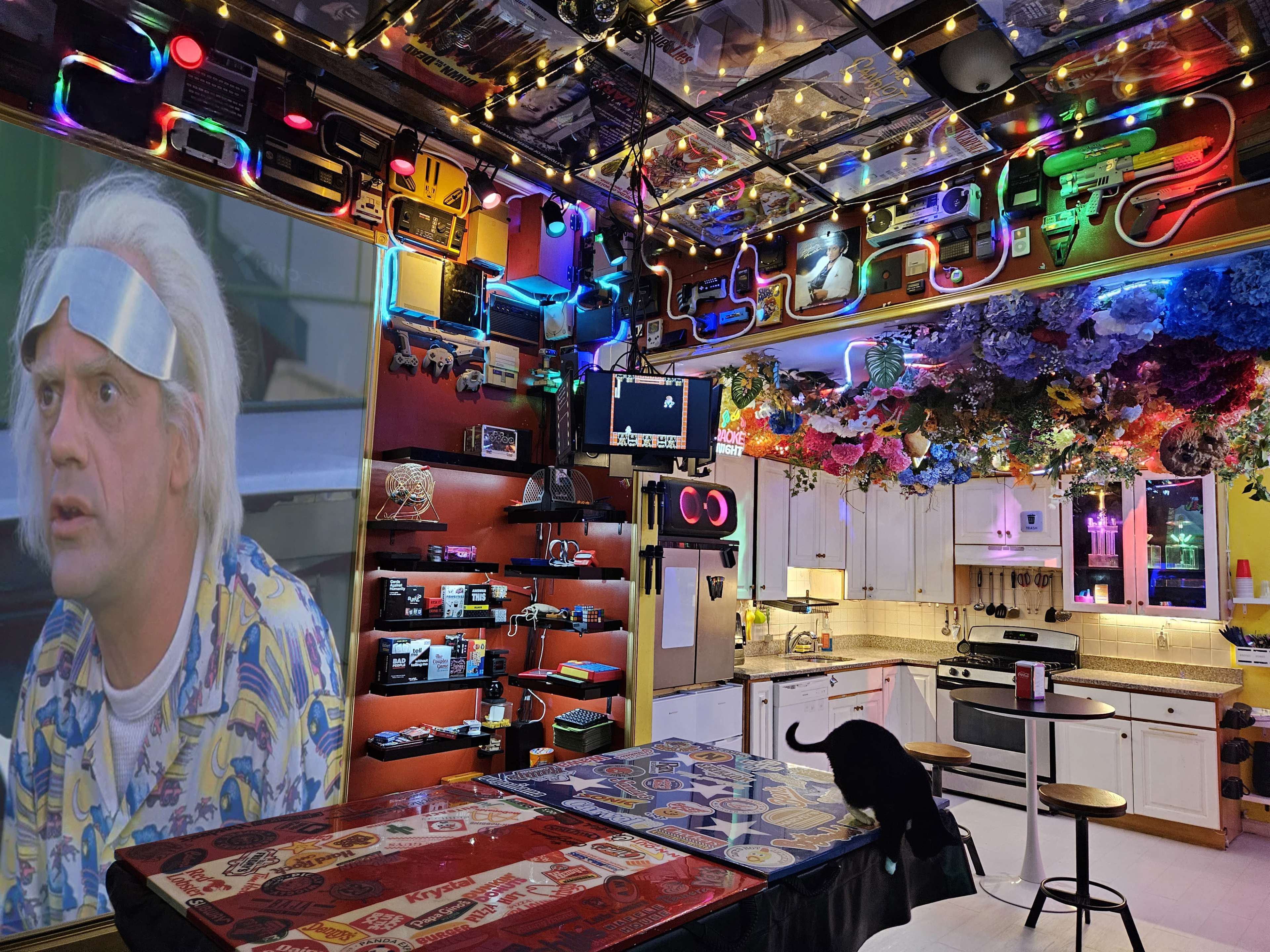 The image shows a colorful, eclectic kitchen with various decorations, bright lights, and a large screen displaying a character from a film.