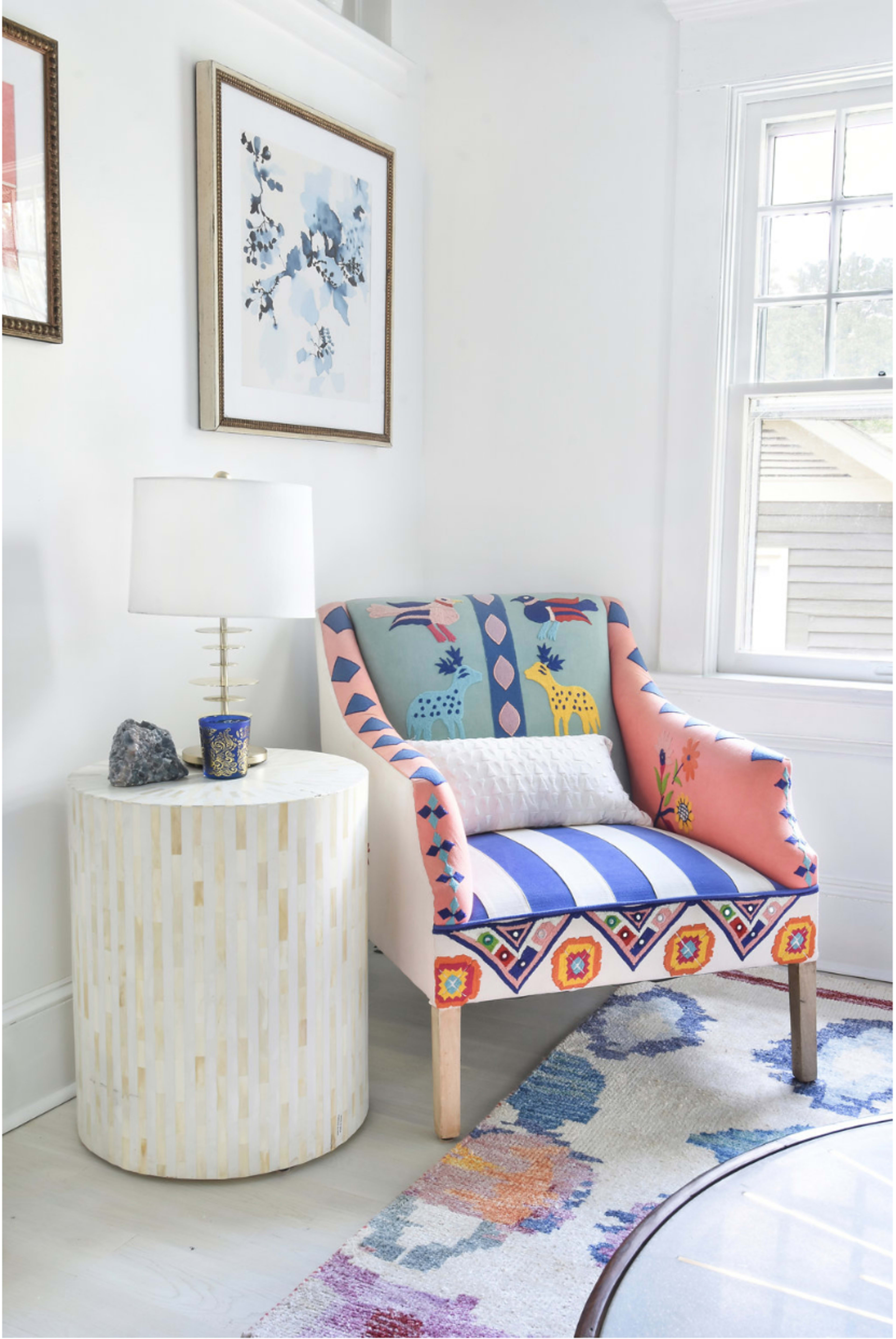 A colorful upholstered armchair is positioned beside a round side table with a lamp and decor in a bright, well-lit room.