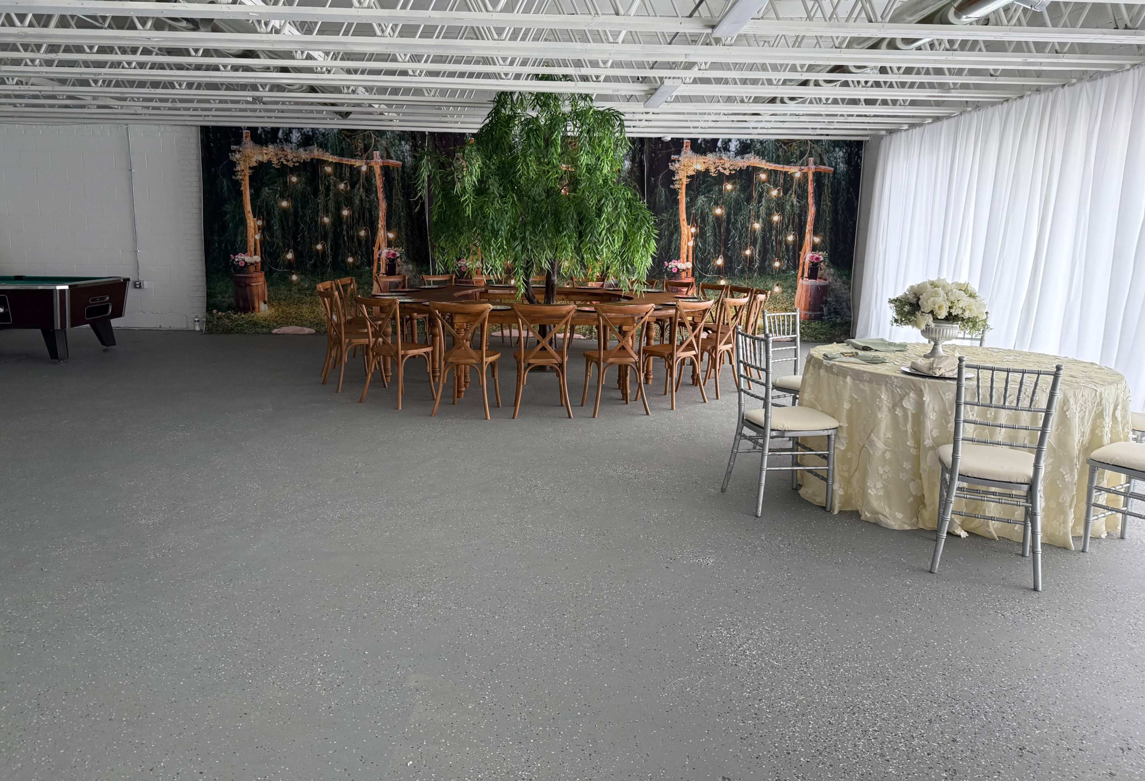 The image shows a large room with a circular dining table set for an event, surrounded by wooden chairs, and a separate table with a light-colored tablecloth, set against a backdrop of artificial greenery and lights.