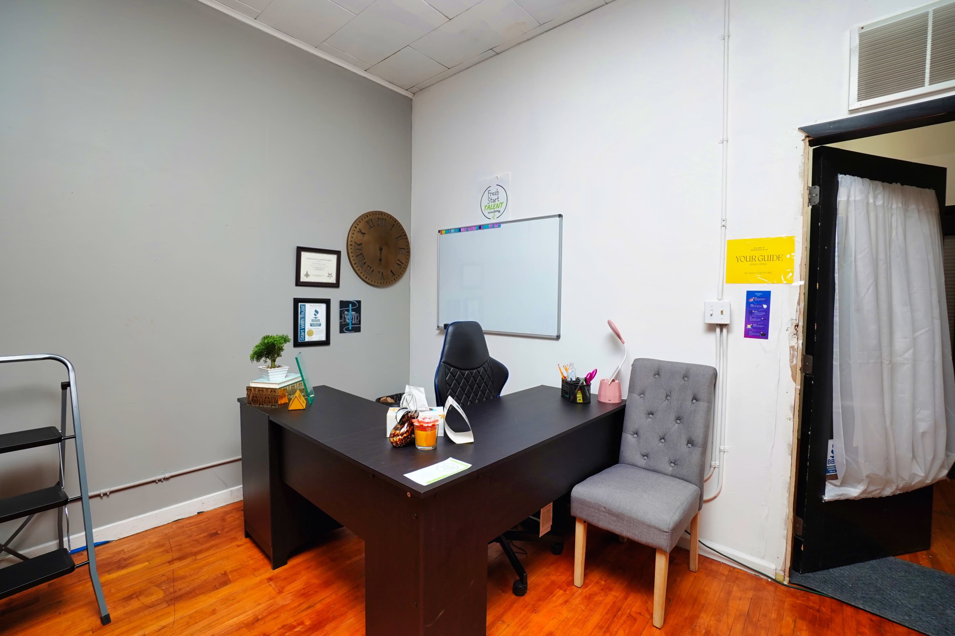 A small office space with a black desk, an office chair, a gray upholstered chair, a whiteboard on the wall, and various decorations including framed certificates and a plant.