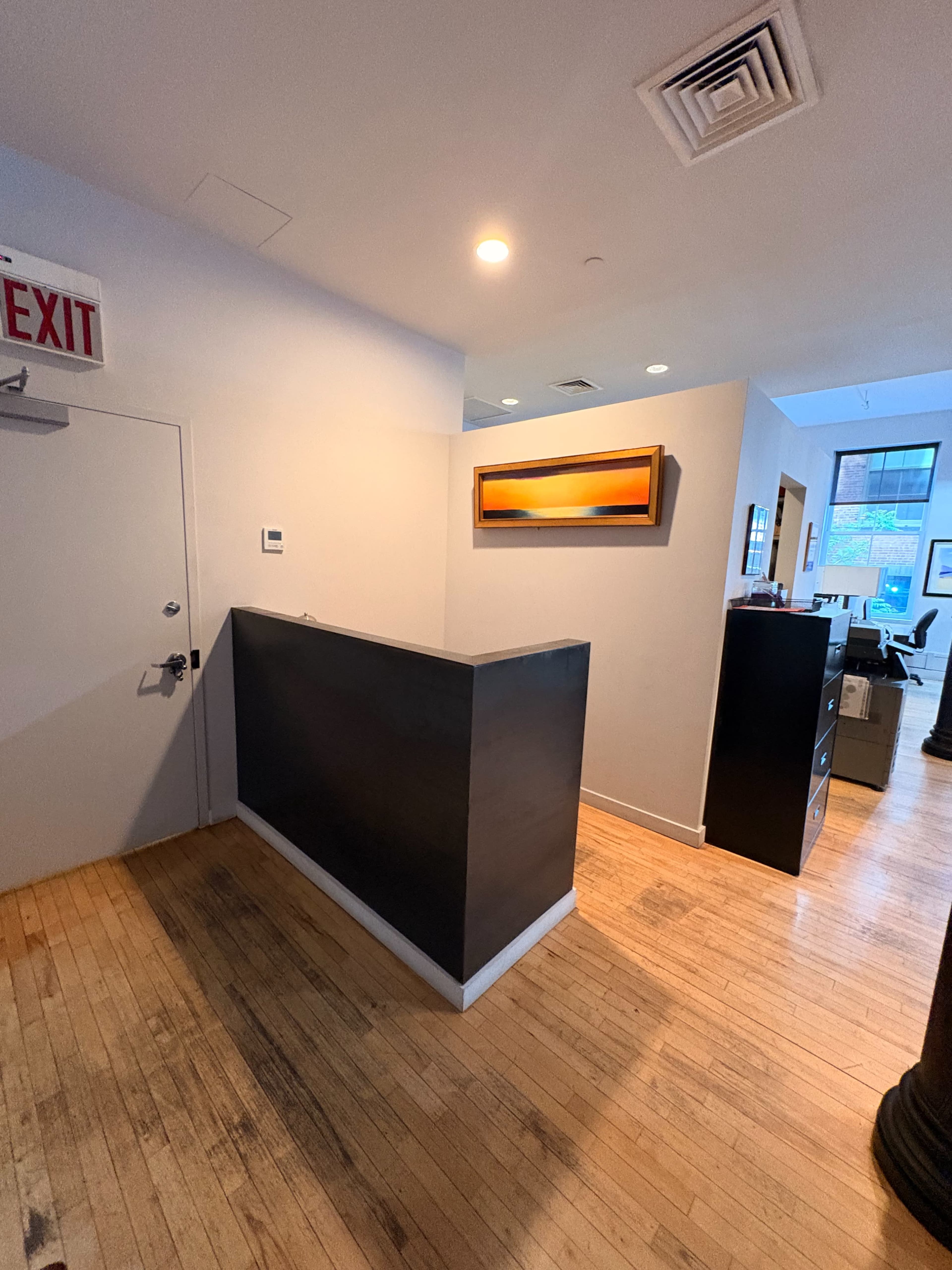 The image shows a modern office reception area with a dark counter, an exit sign, and a wall-mounted artwork, leading into an office space with wooden flooring.