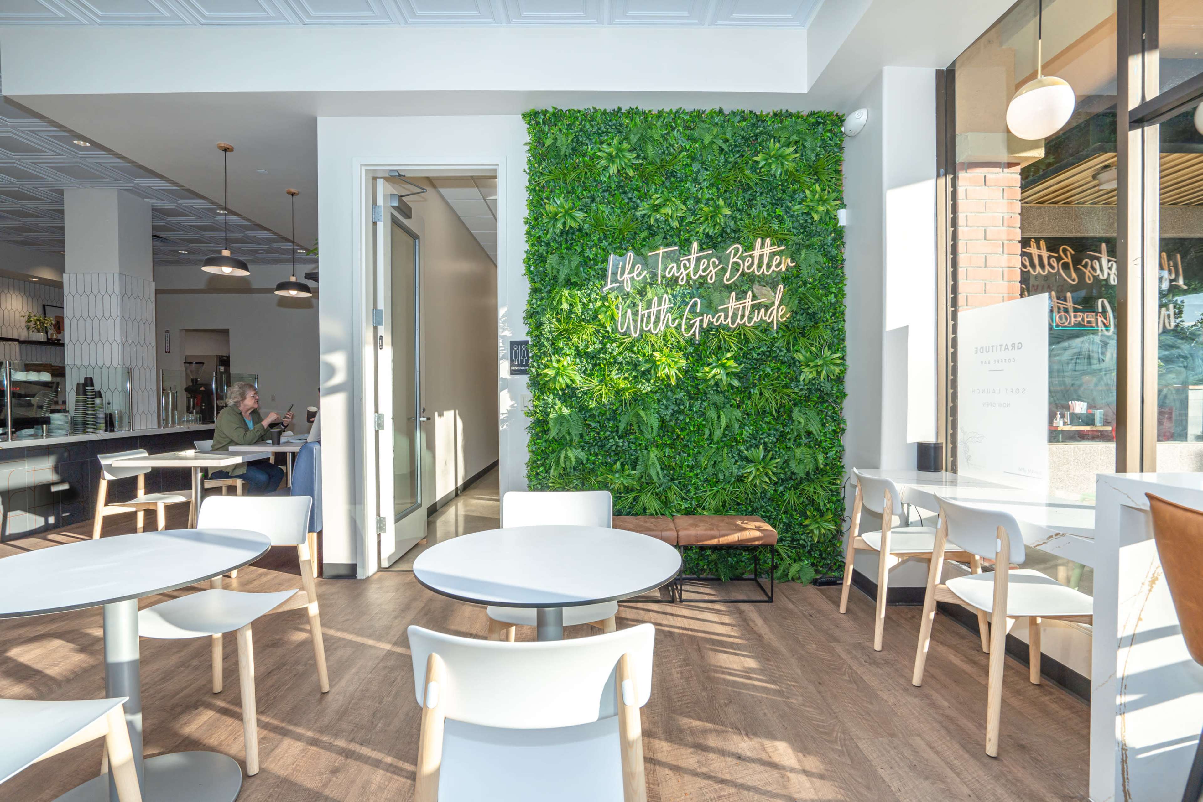 A modern café interior features a green wall with the text "Life Tastes Better With Gratitude," white tables, and light-colored chairs.