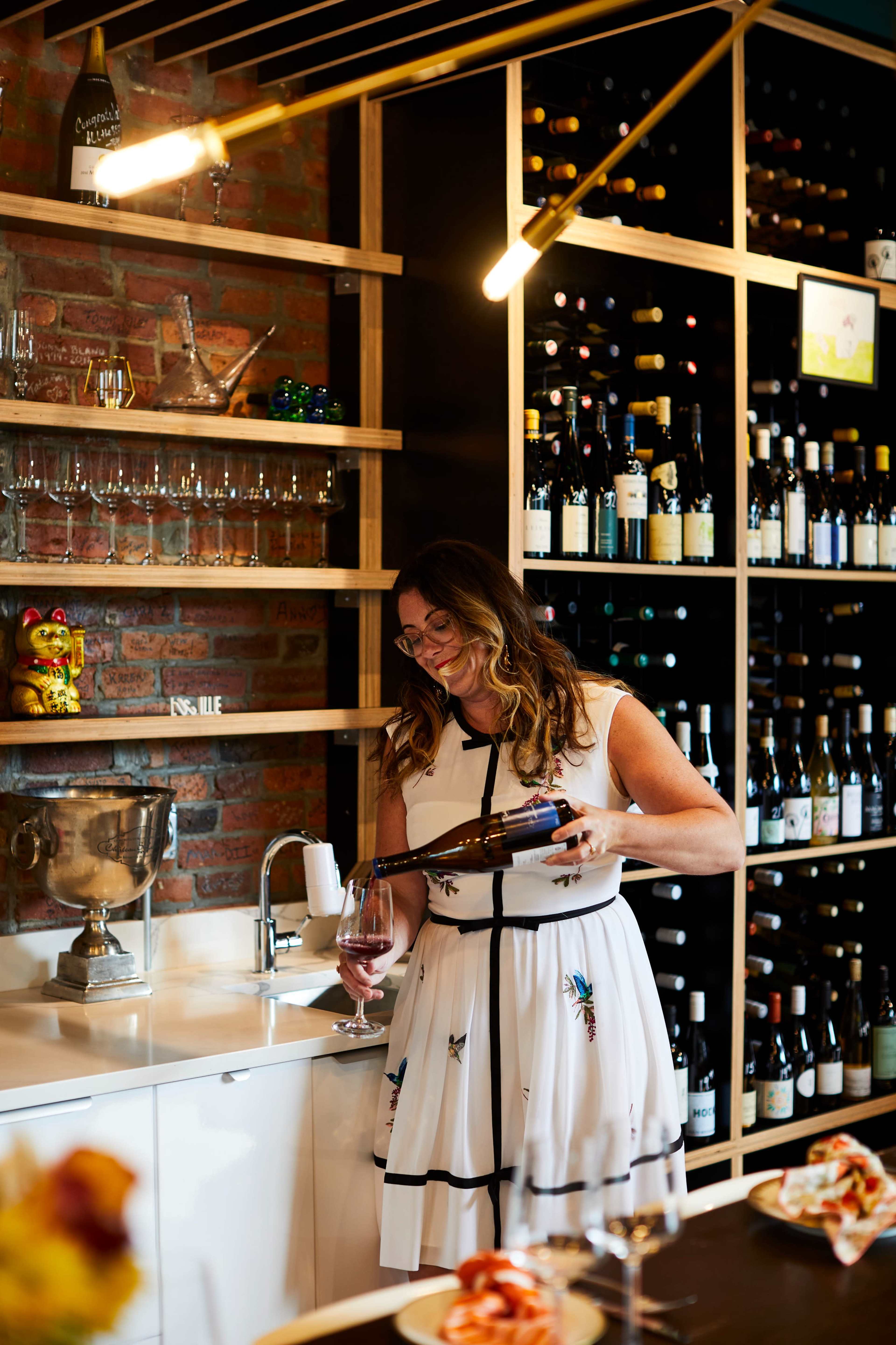 A woman in a white dress pours wine into a glass in a wine bar with shelves of bottles in the background.