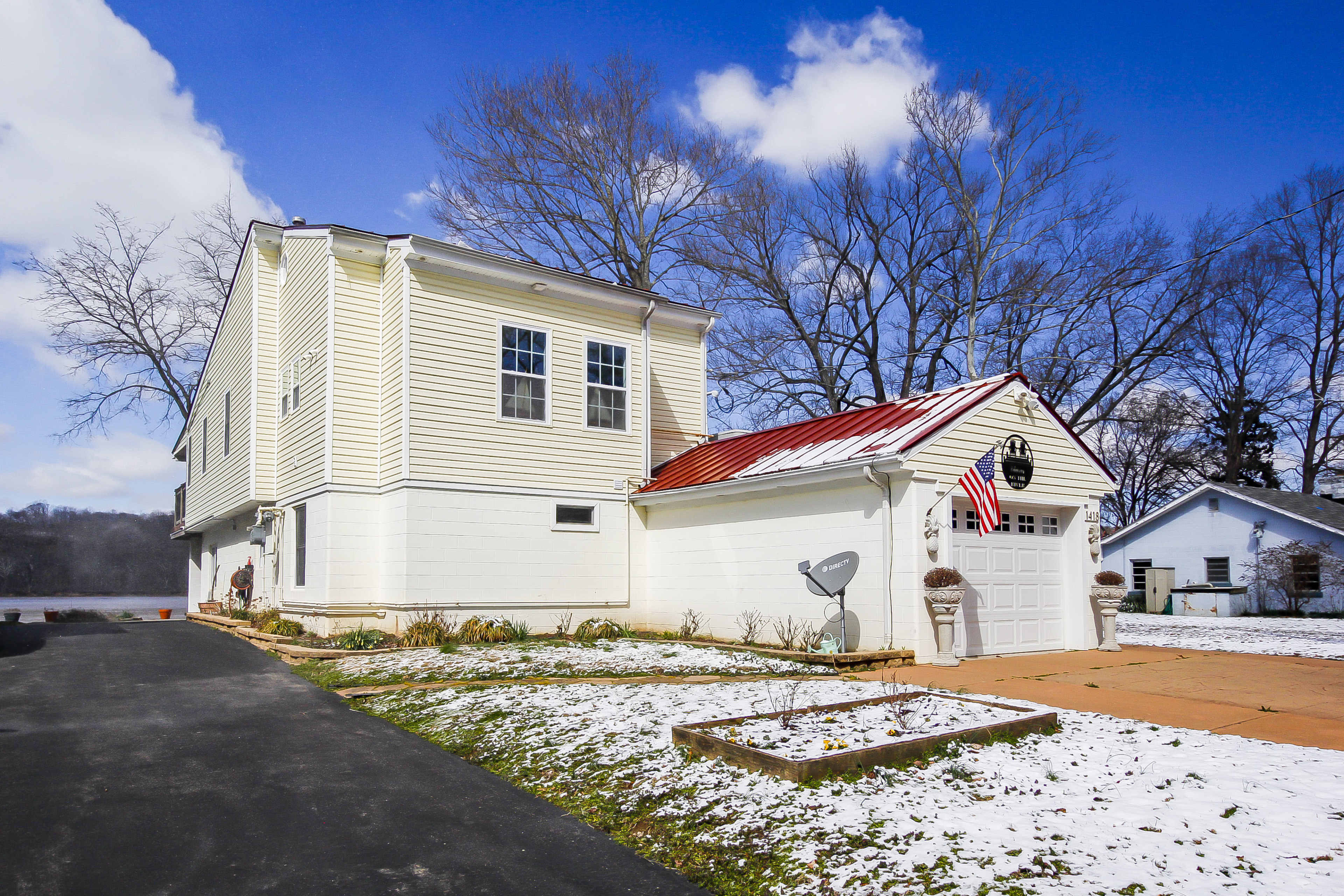 A two-story house with a red metal roof garage, situated on a snowy landscape under a clear blue sky.