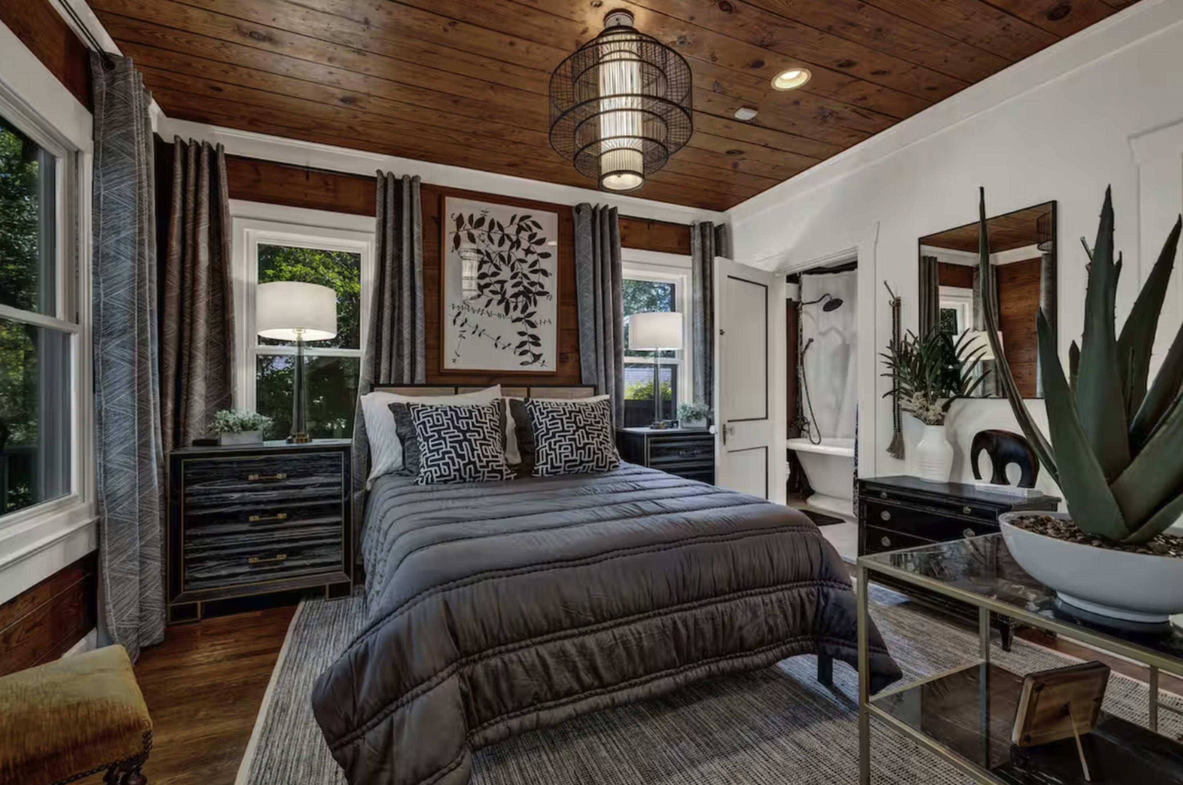 The image shows a modern bedroom featuring a bed with gray bedding, wooden walls, large windows with curtains, and decorative elements like plants and artwork.