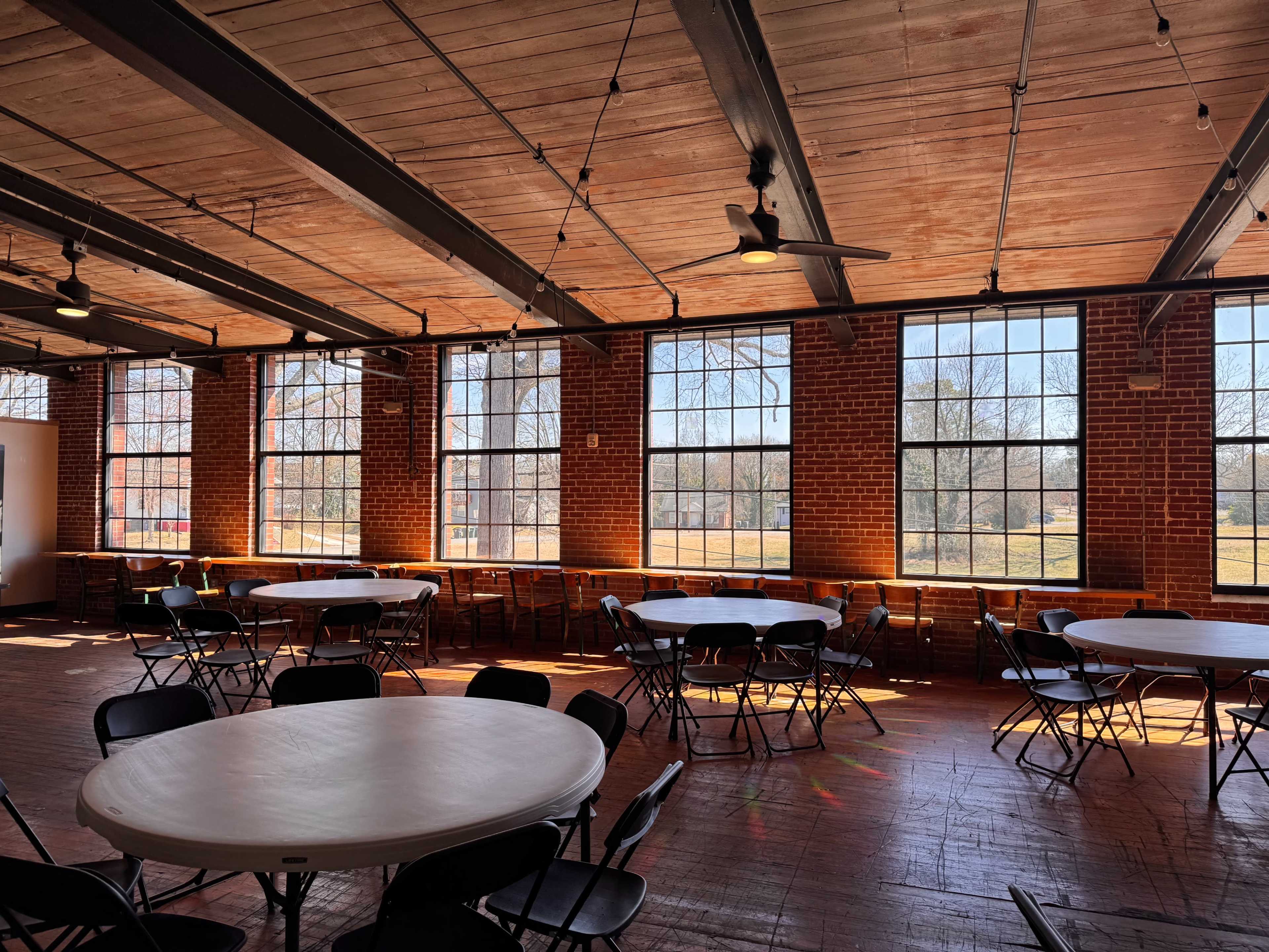 The image shows an indoor space with several round tables and black chairs, characterized by large windows that allow sunlight to fill the room.