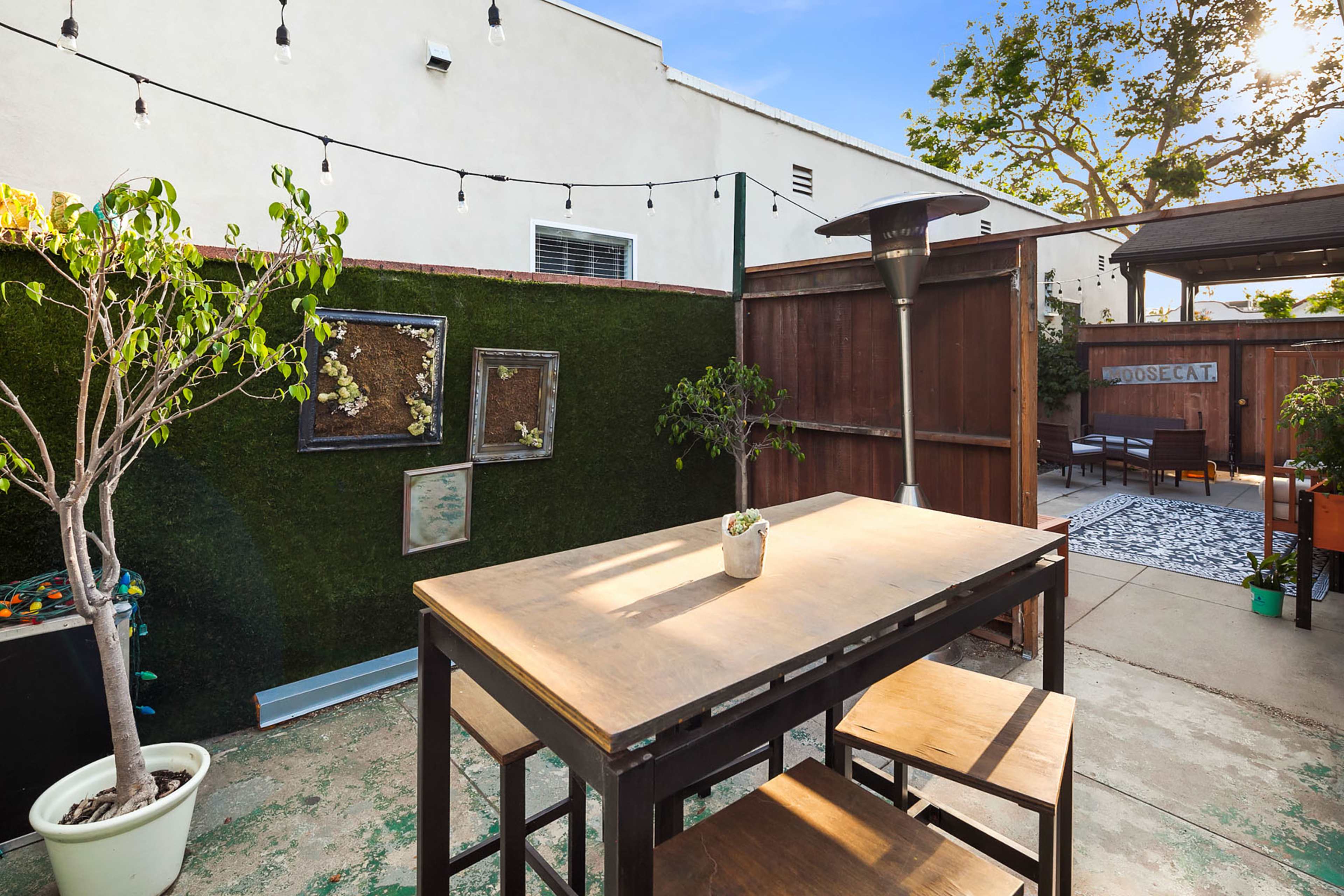 A small outdoor patio features a wooden table and chairs, surrounded by greenery and decorative elements, with a gas heater and string lights.