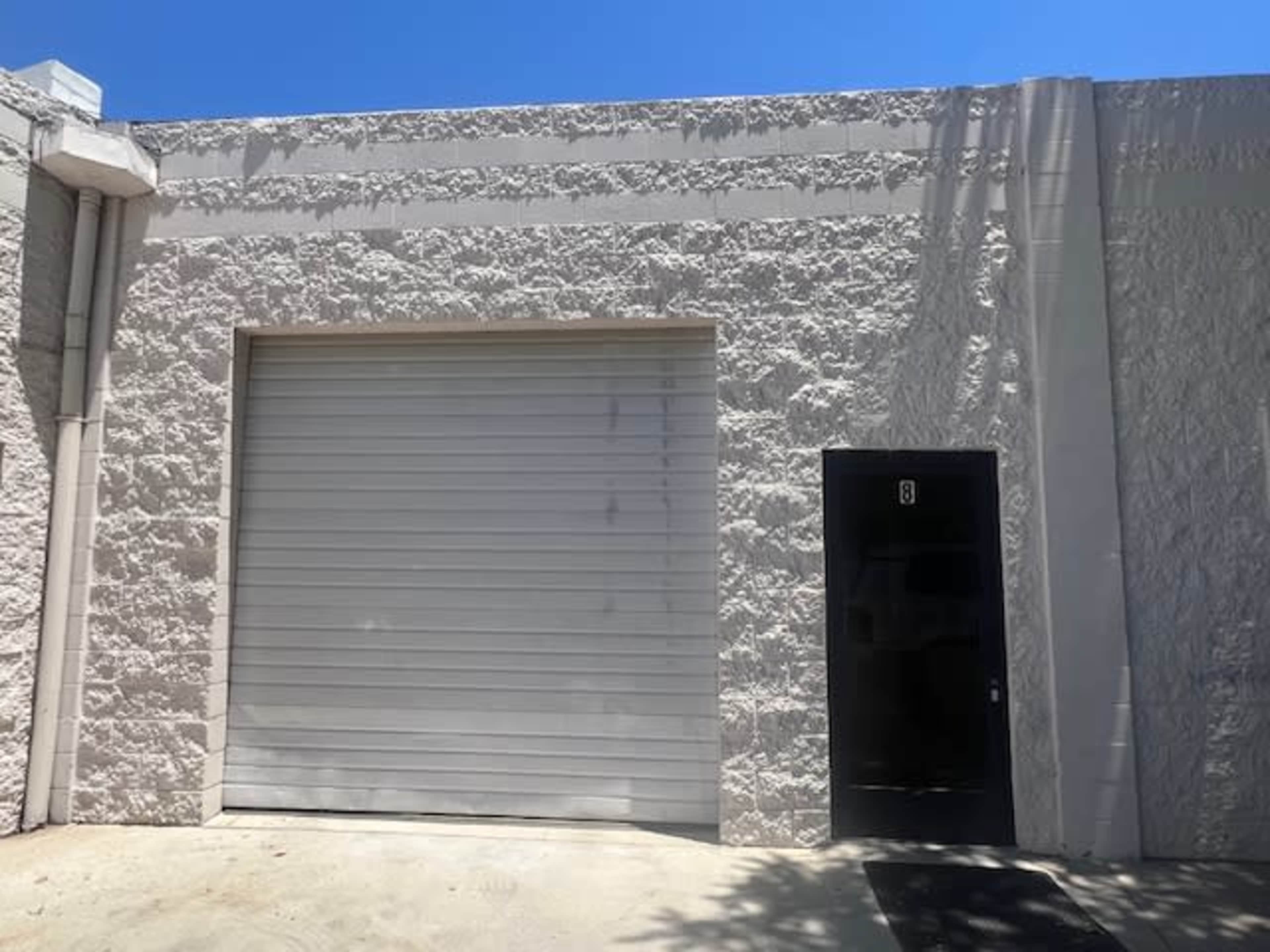 The image shows a light-colored building exterior with a large, closed roll-up garage door and a black door to the right.