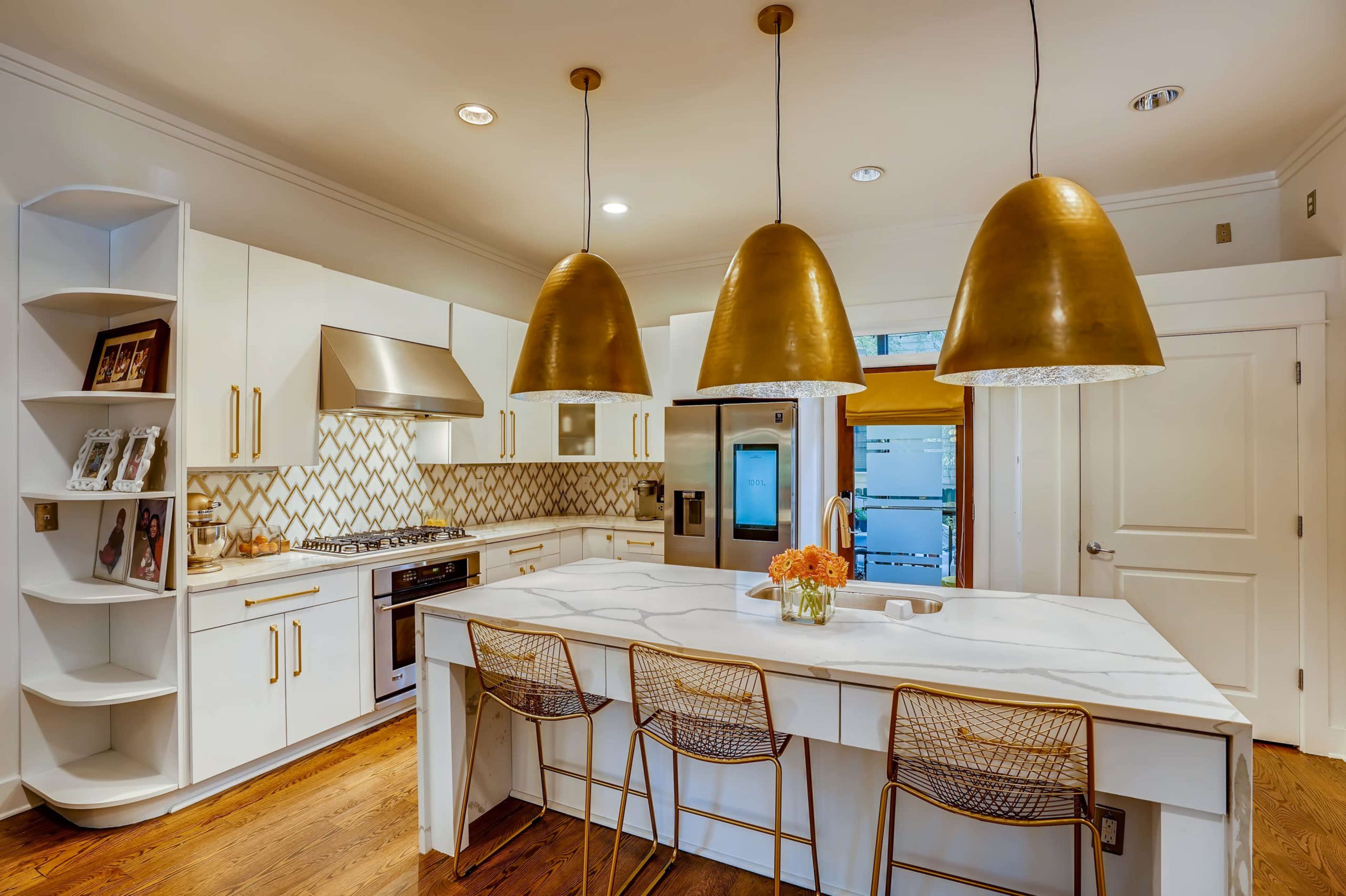 A modern kitchen features white cabinets, a marble island with three gold pendant lights overhead, and a decorative tile backsplash.