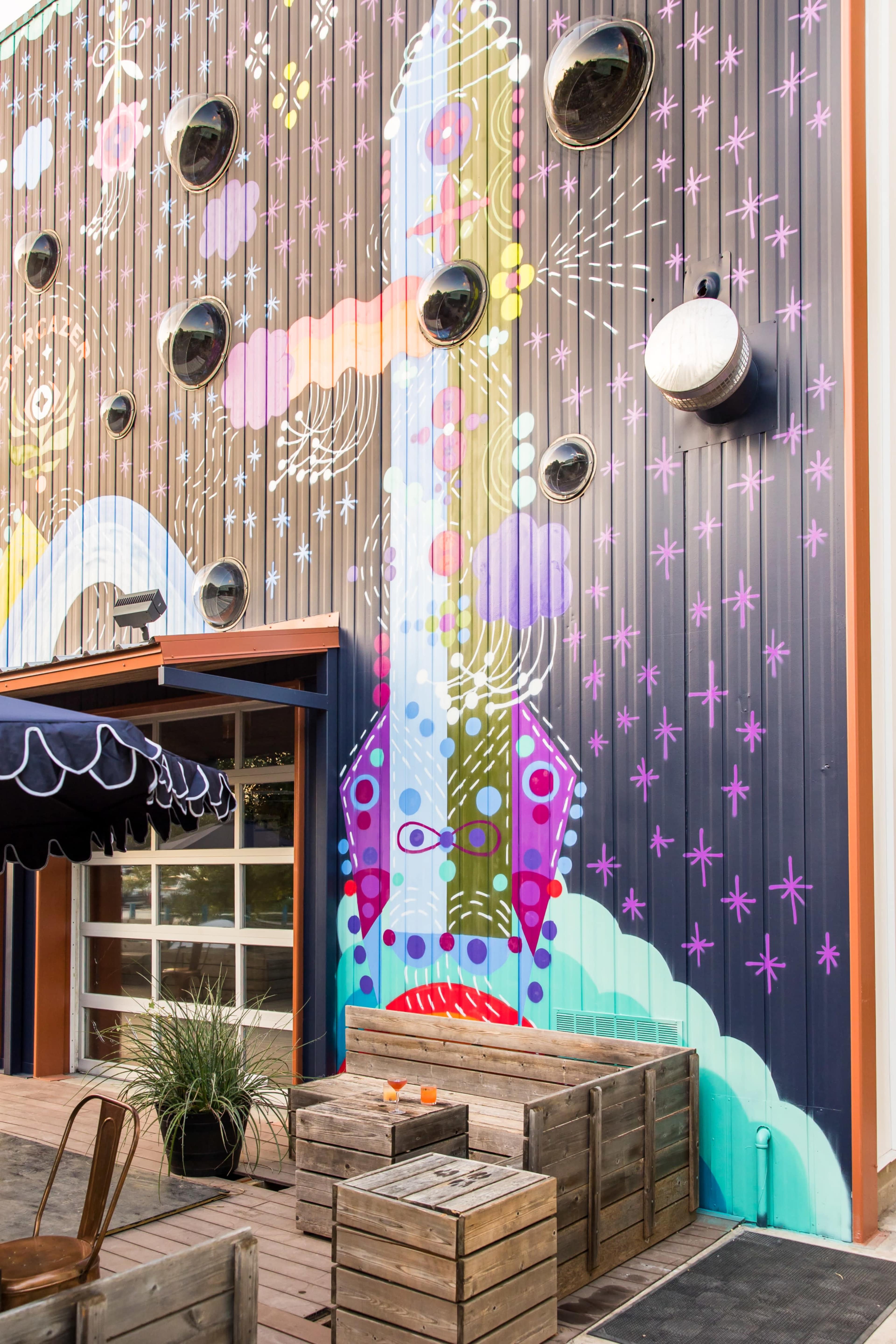 A colorful mural depicting a guitar adorns the side of a building, with wooden seating arranged on a patio in front.