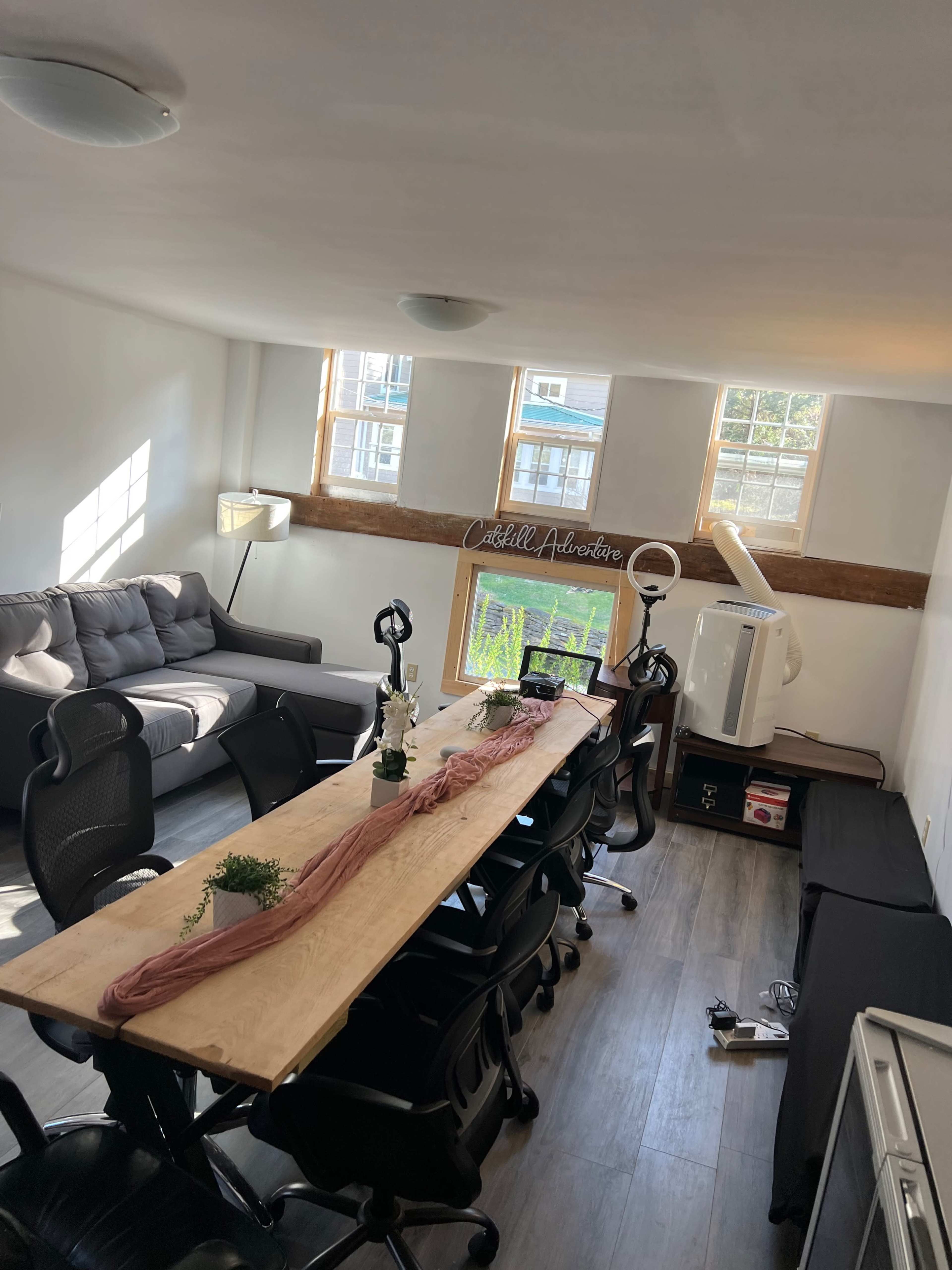 A bright office space featuring a long wooden table surrounded by black office chairs, a gray couch, large windows, and a decorative sign on the wall.