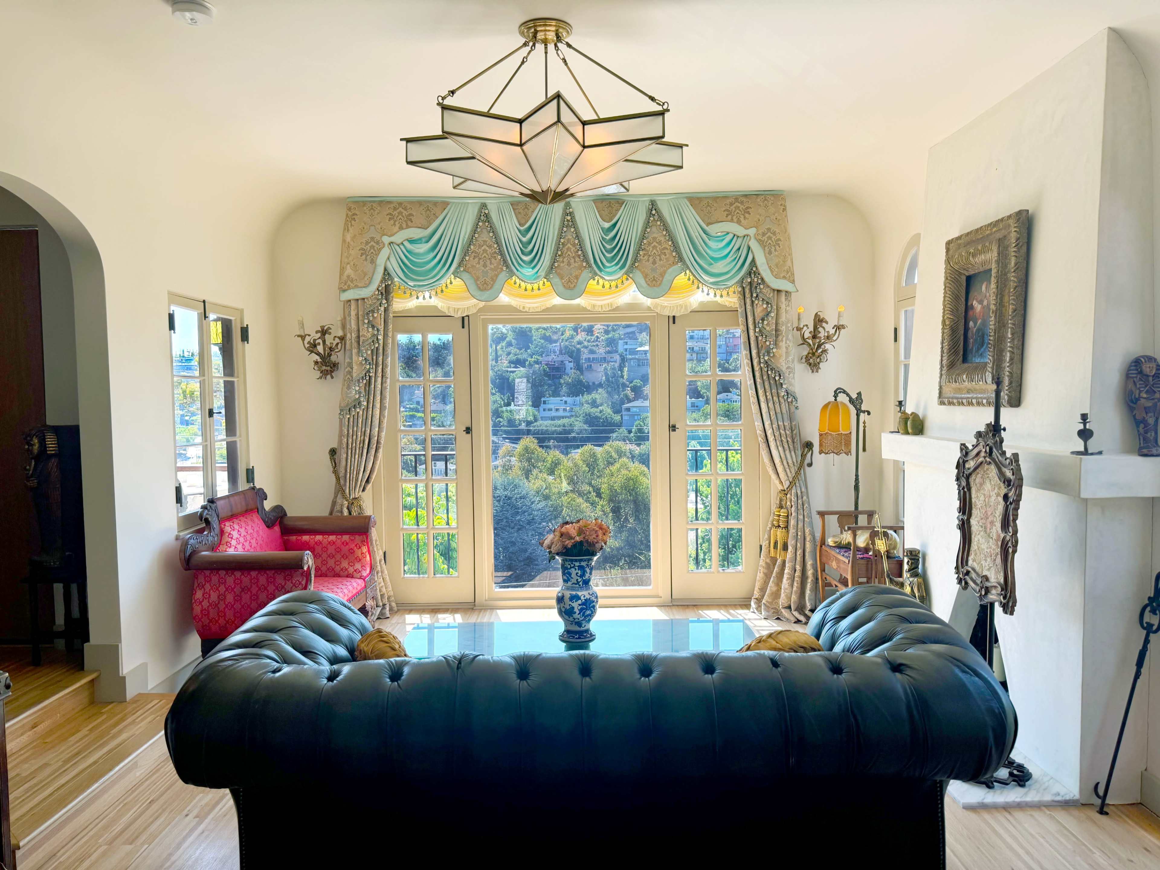 A living room with a black leather chesterfield sofa, large windows with turquoise drapes, and a view of a green landscape.