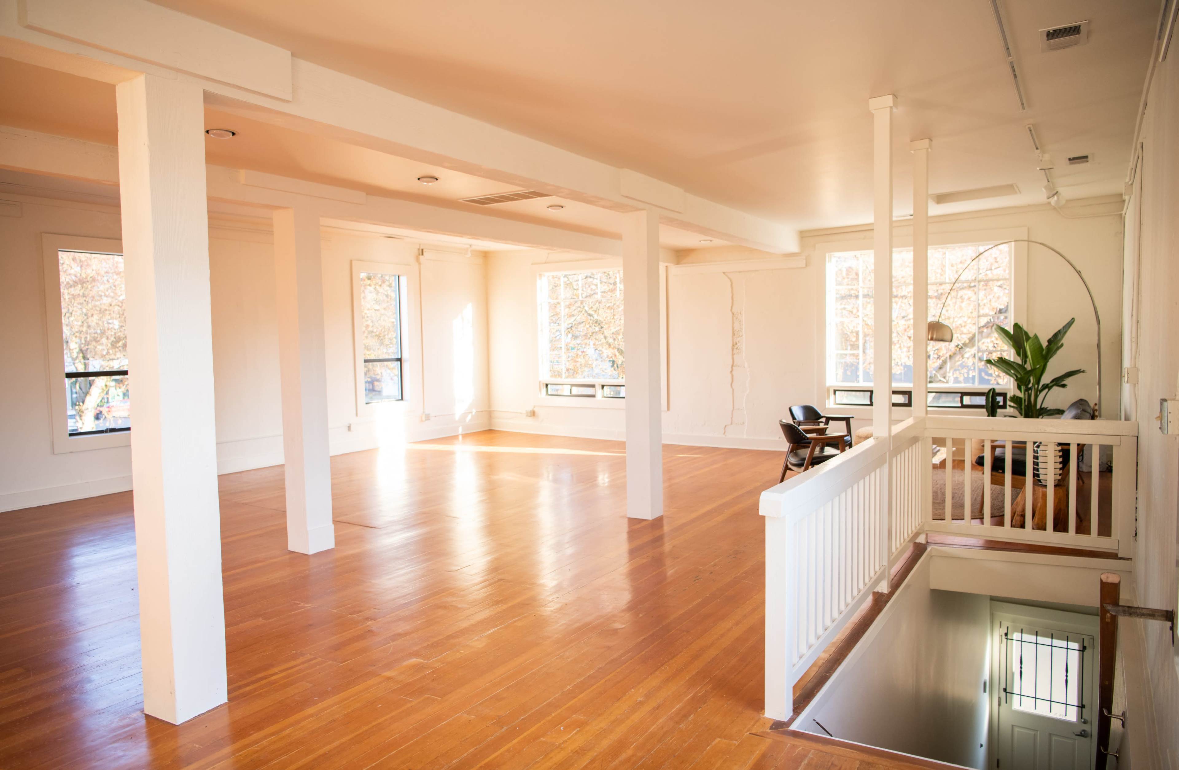 A spacious, empty room with wooden flooring, large windows, and white walls, featuring a staircase leading to a lower level.