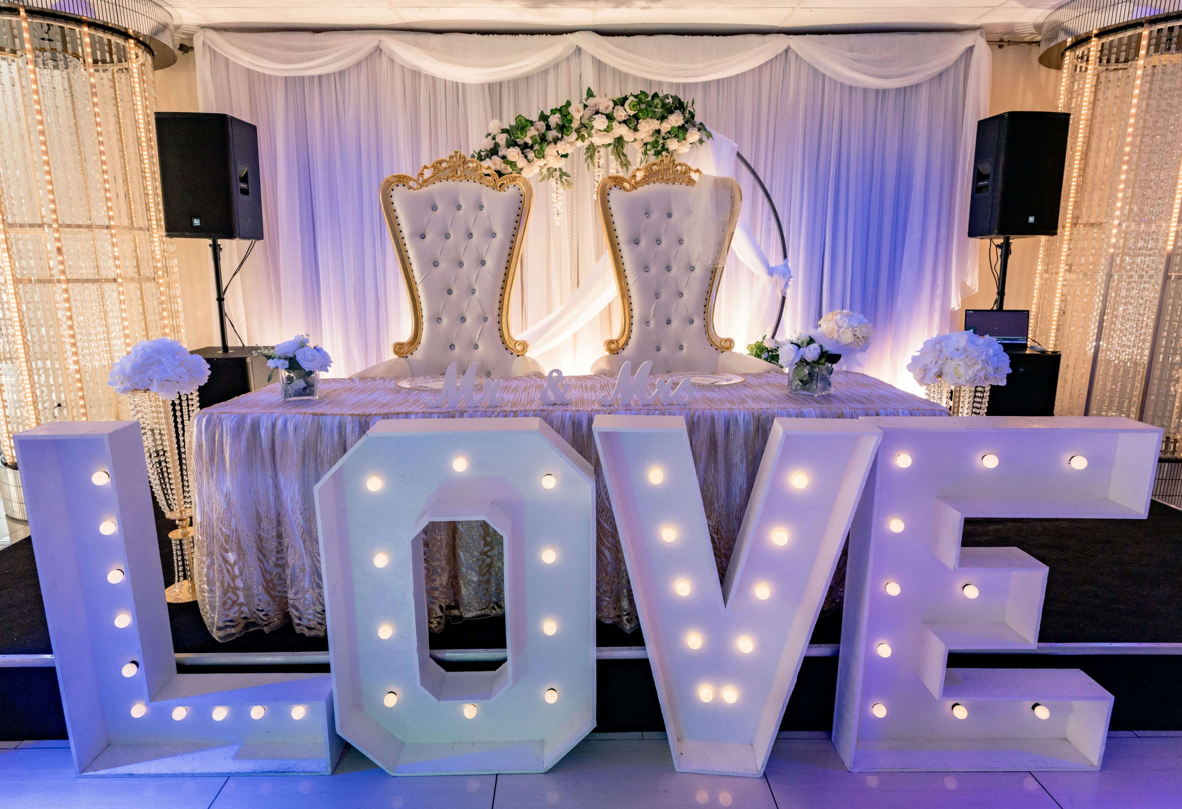 The image features a wedding reception setup with two elegant chairs at the center, adorned with floral arrangements, and a large illuminated "LOVE" sign in front.